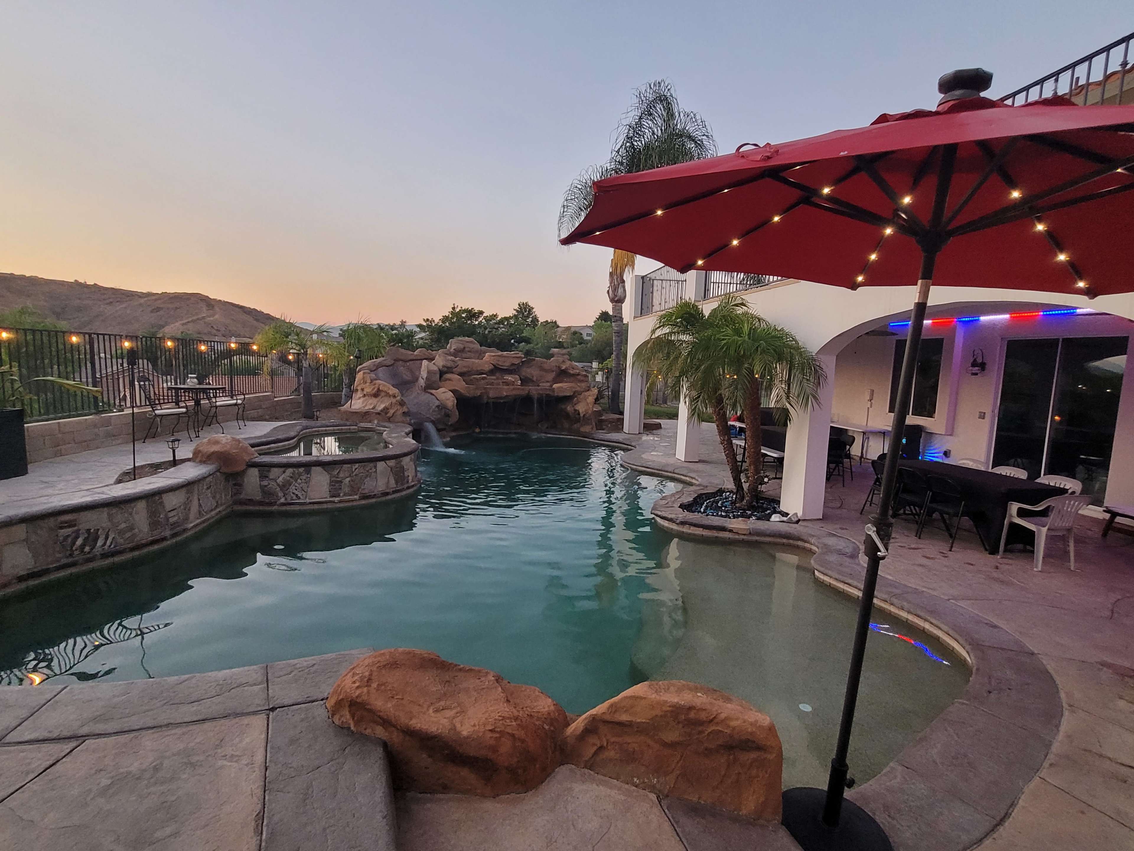 A backyard pool area features a stone-decked pool with a waterfall, surrounded by palm trees and lounge furniture, under a red umbrella at sunset.