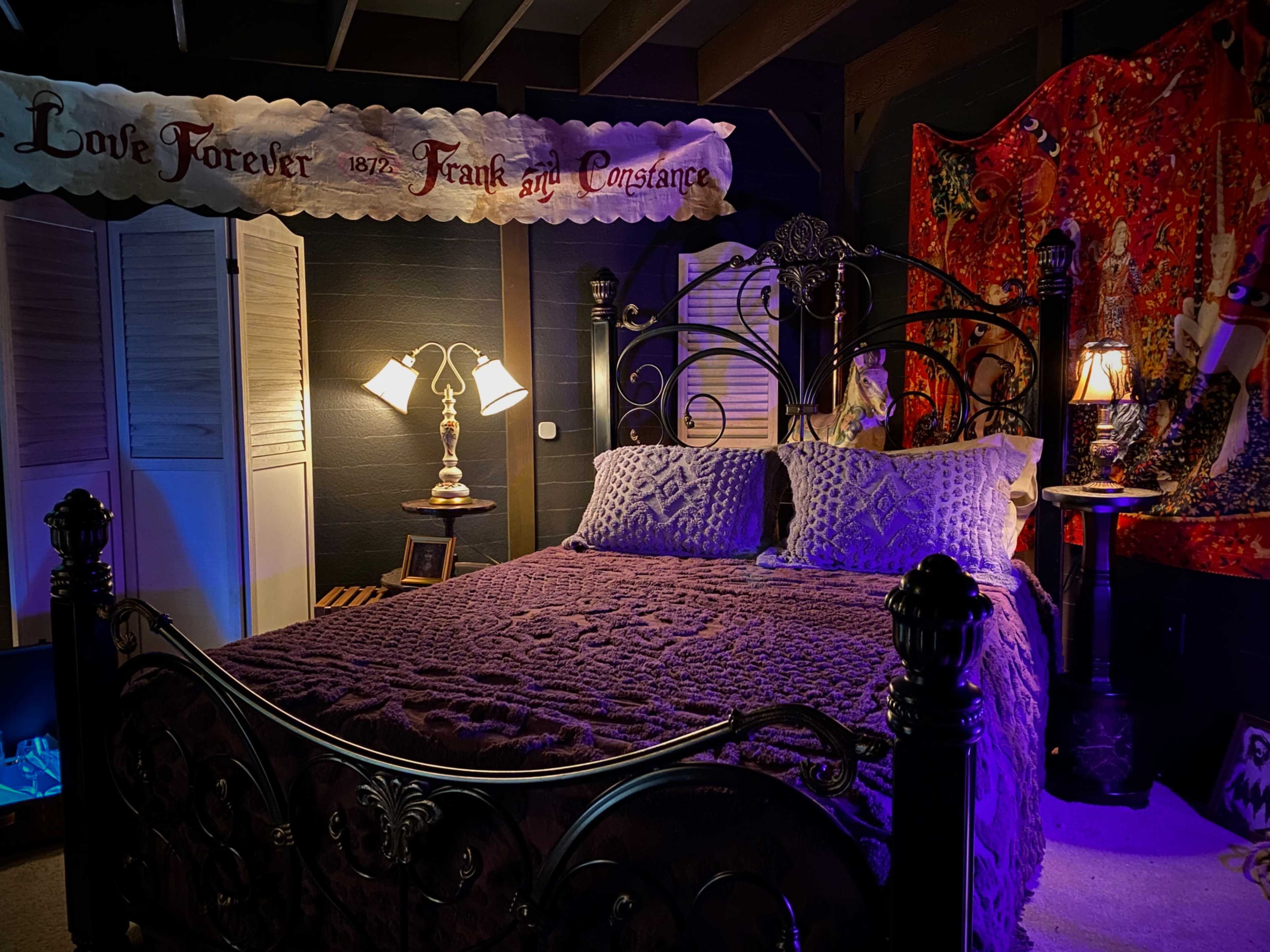 The image shows a dimly lit bedroom featuring a wrought iron bed with a purple bedspread, decorative pillows, and a patterned wall hanging.