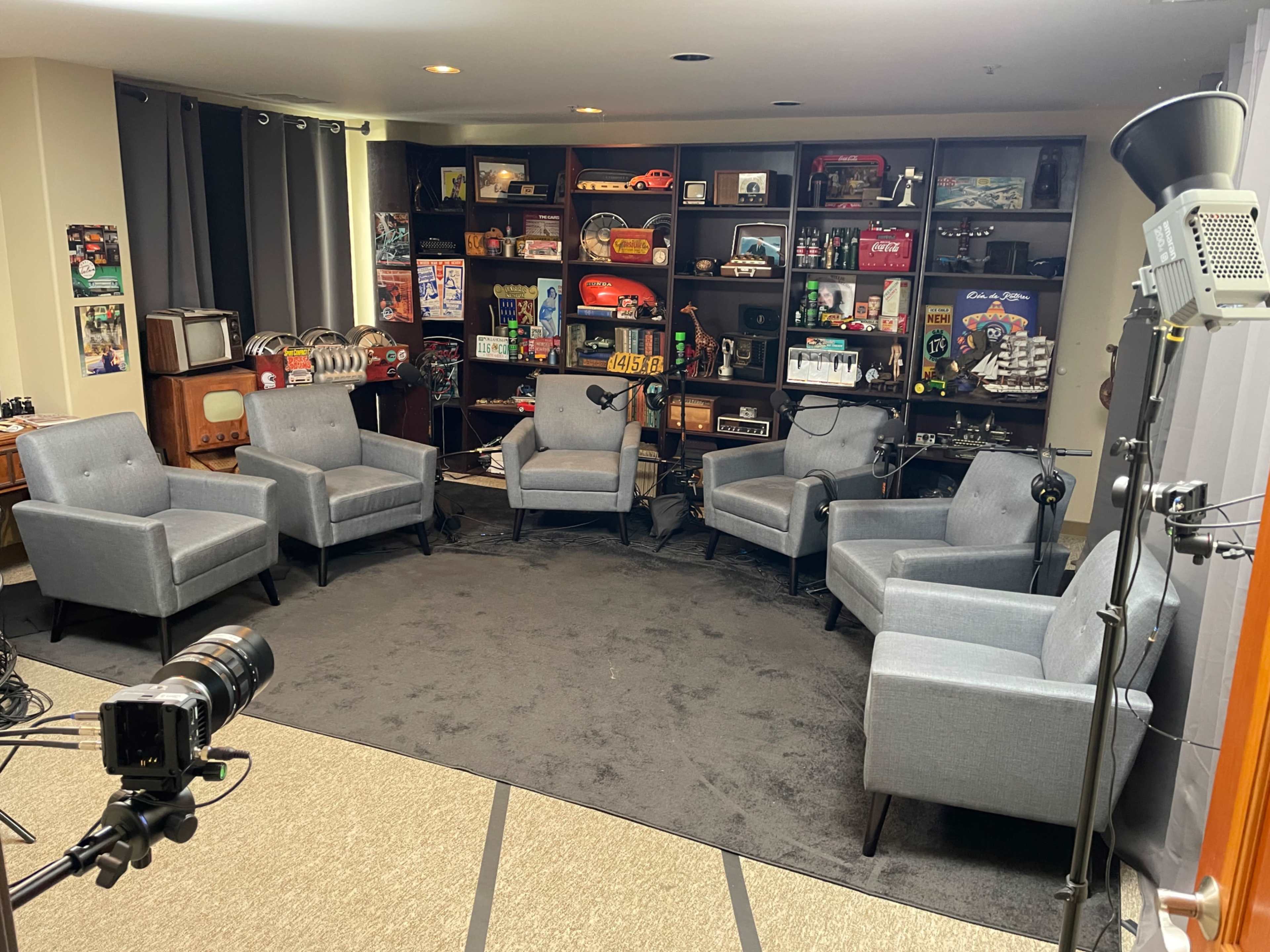 The image shows a cozy meeting space with six gray armchairs arranged in a circle on a dark carpet, surrounded by shelves filled with various vintage items and memorabilia.