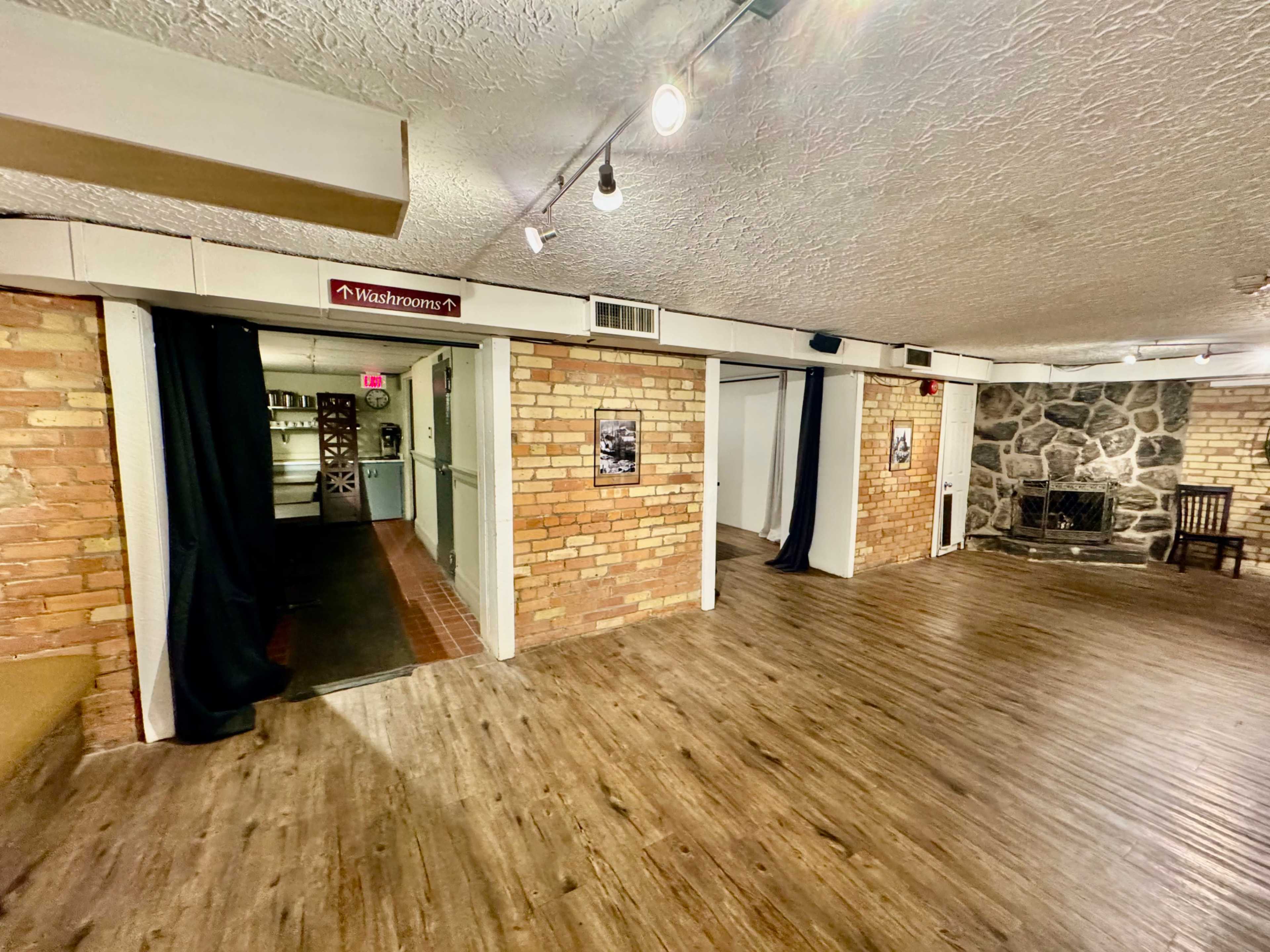 A spacious interior room with brick walls, wood flooring, and a stone fireplace, featuring doorways leading to restrooms and a dimly lit area.