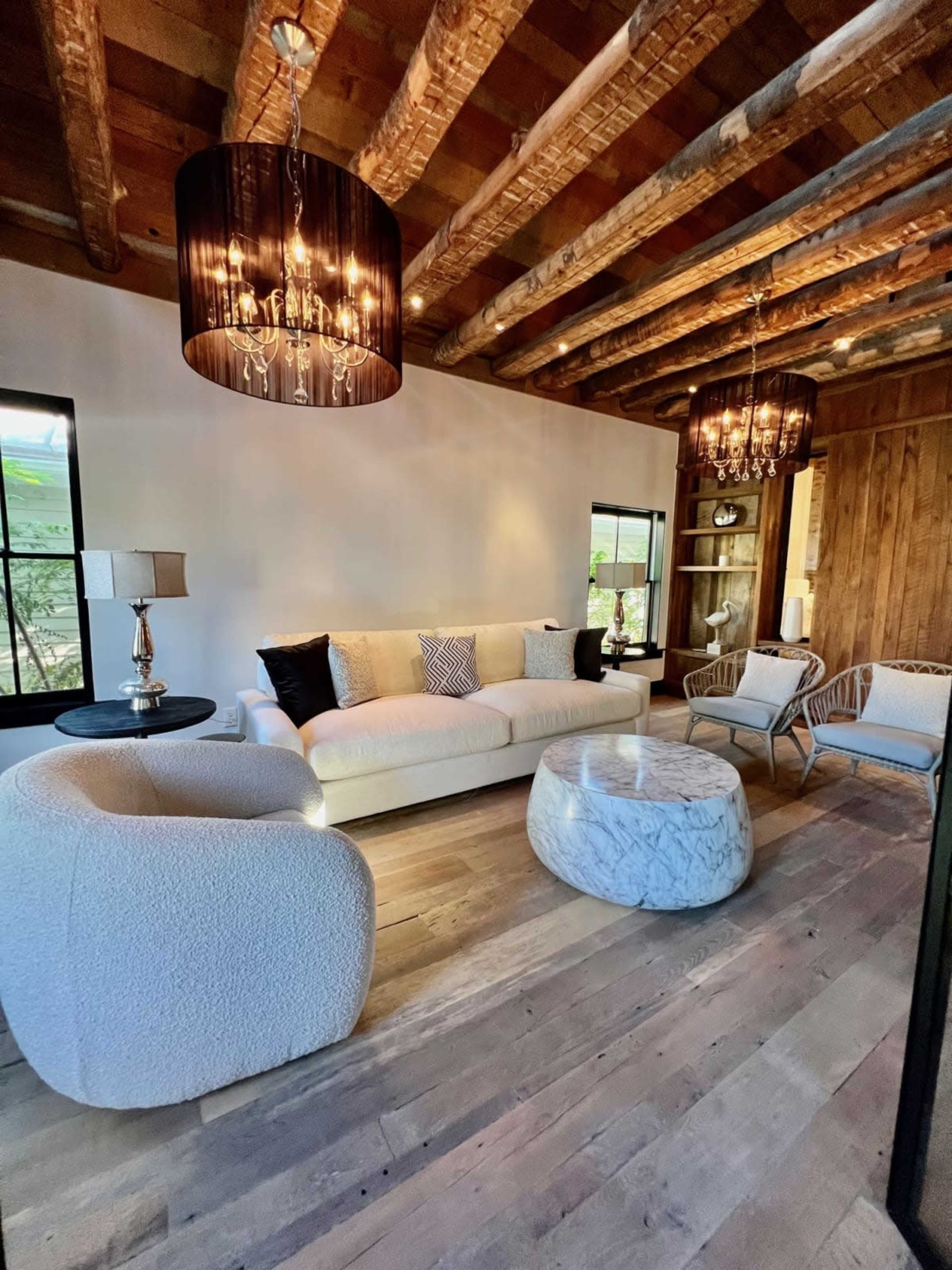 A spacious living room with a large white sofa, two armchairs, a marble coffee table, and wooden ceiling beams, all illuminated by stylish light fixtures.