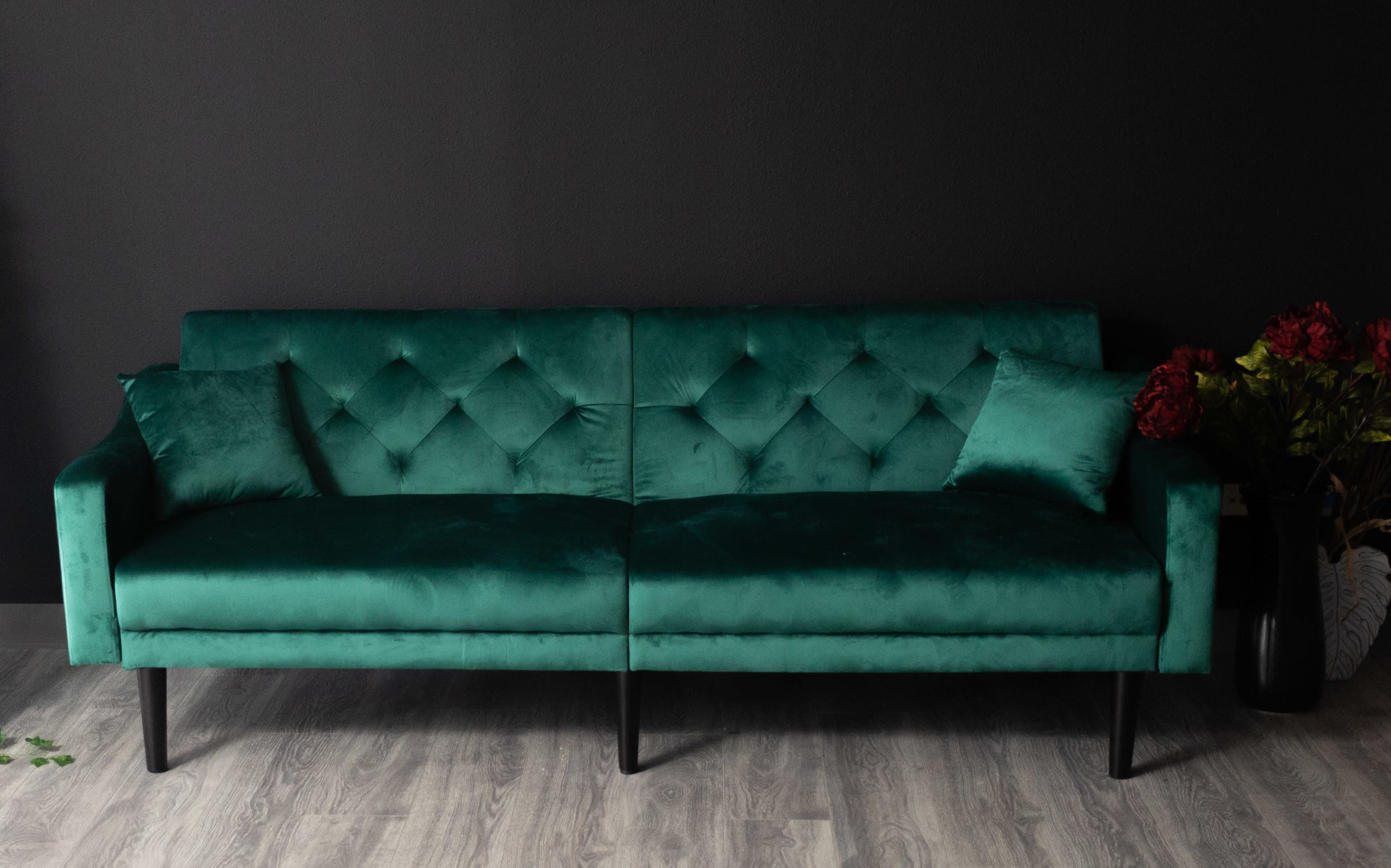 A green velvet sofa with diamond tufting sits against a dark wall, accompanied by a decorative pillow and floral accents.