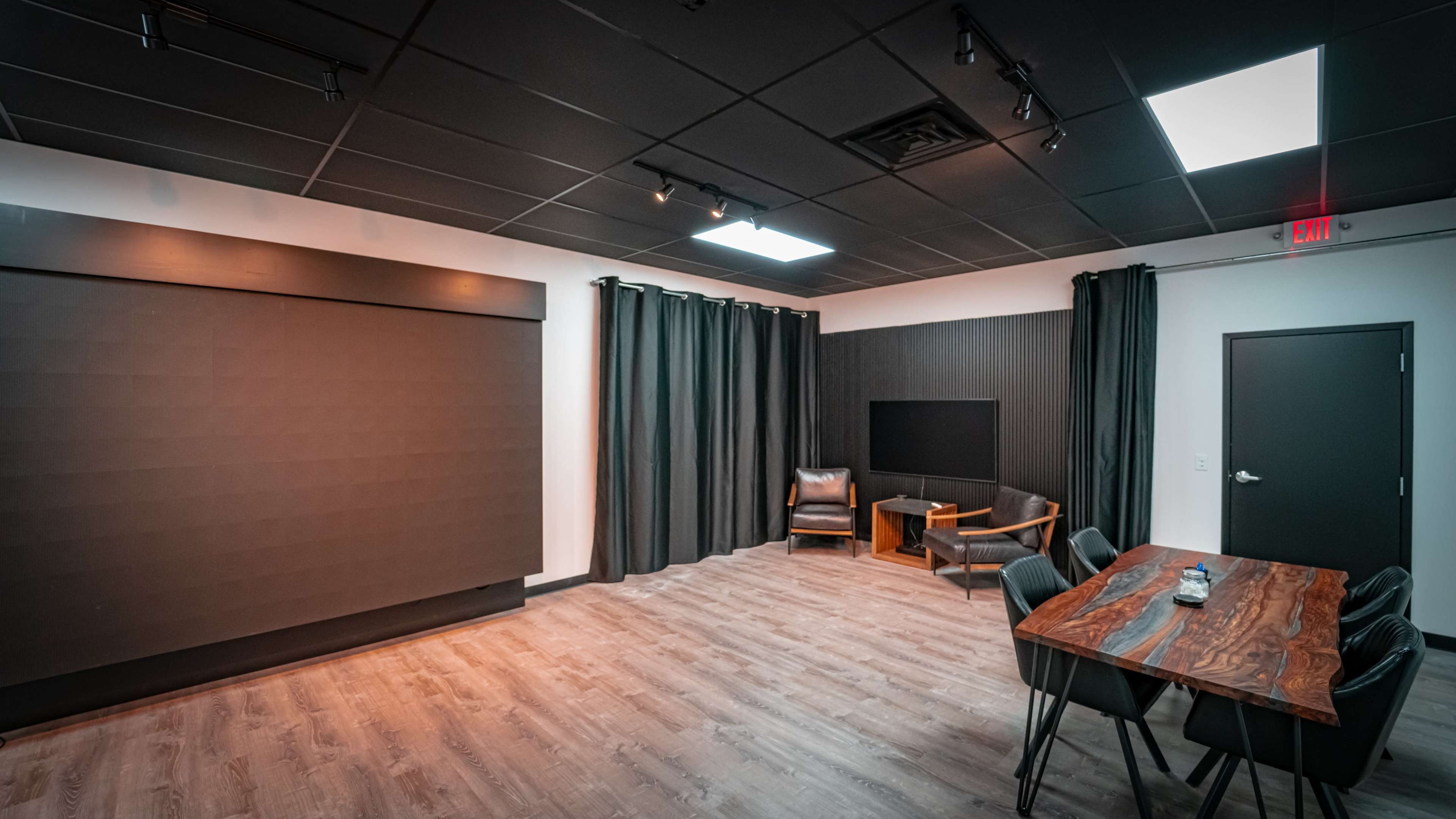 A modern room with a dark color scheme, featuring a large wall screen, a seating area with two chairs, and a wooden table surrounded by black chairs.