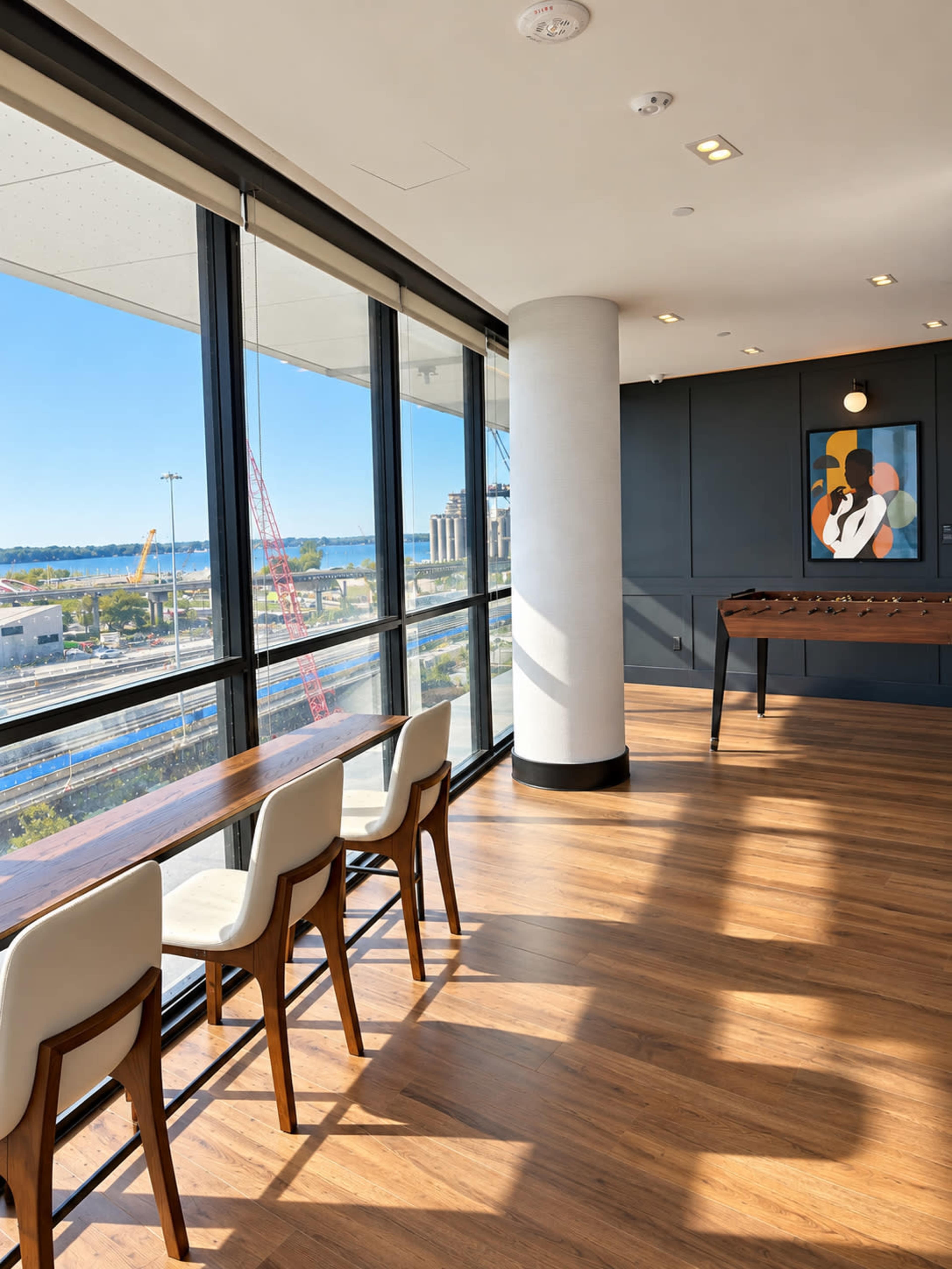 The image shows a modern lounge area with large windows overlooking a construction site and water, featuring wooden flooring and a foosball table.