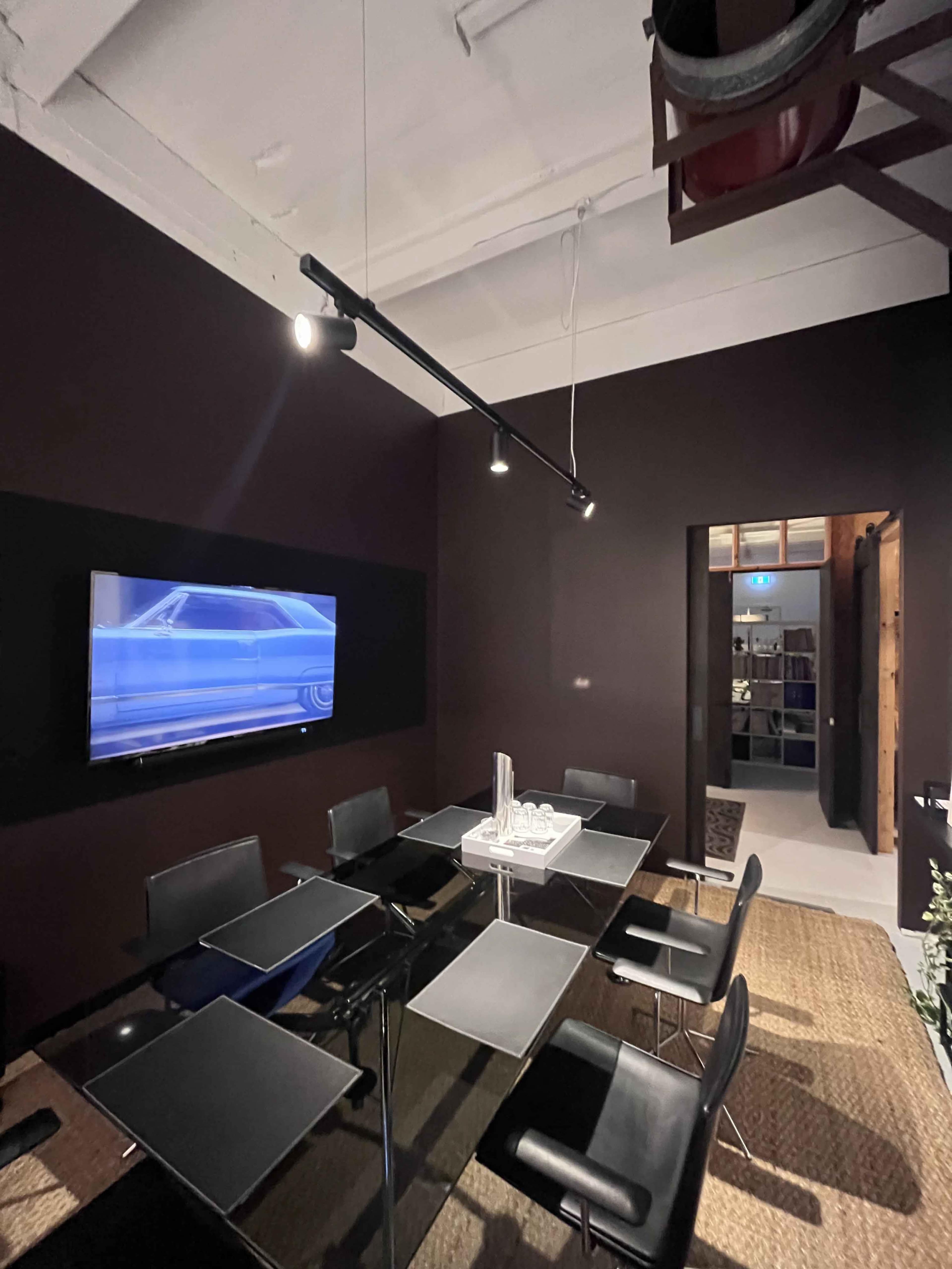 A modern conference room features a glass table surrounded by eight black chairs, with a large screen mounted on the dark wall.