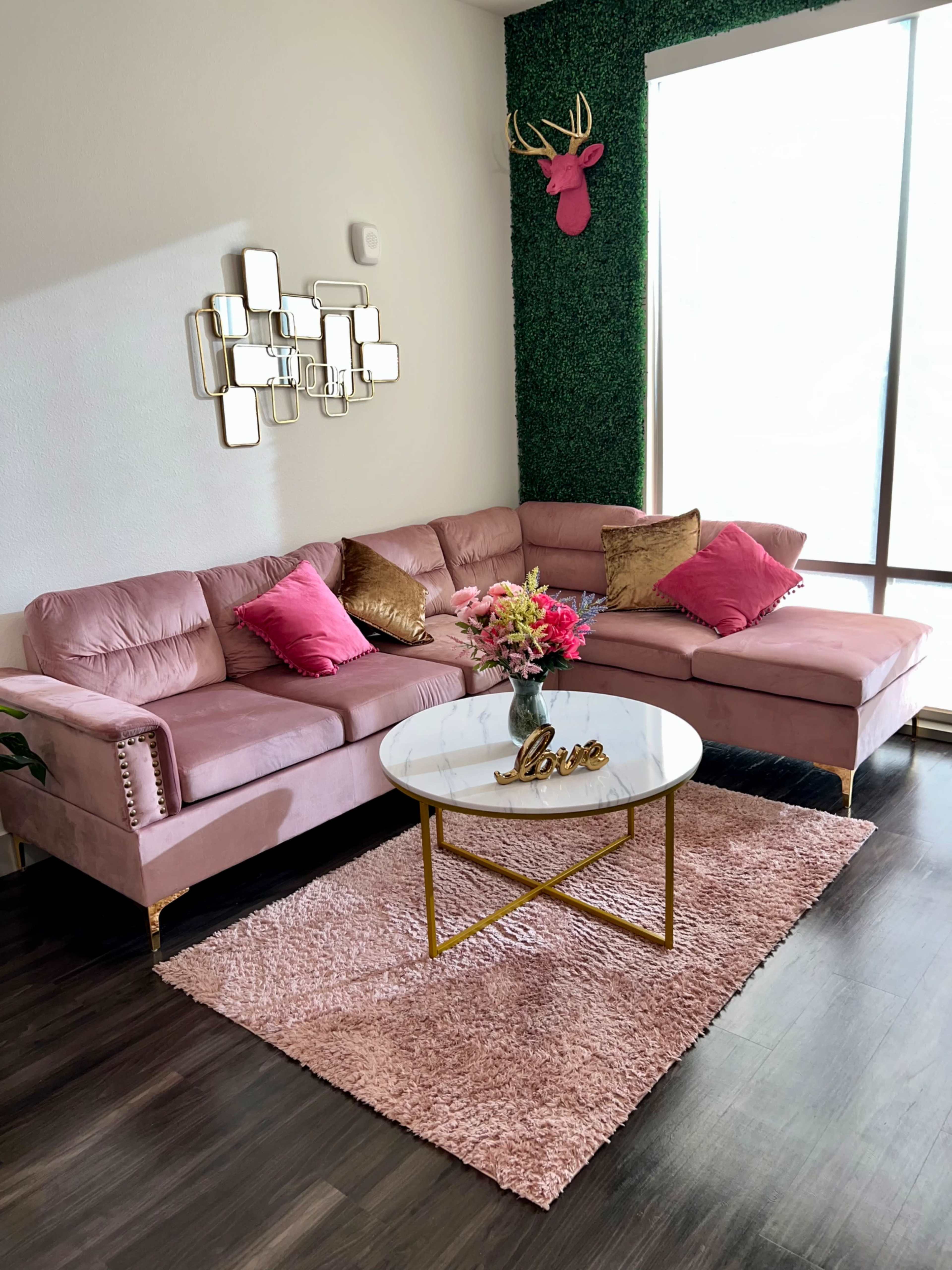 A pink sectional couch with decorative pillows and a glass coffee table sits on a plush rug in a well-lit living room adorned with a green accent wall and wall decor.