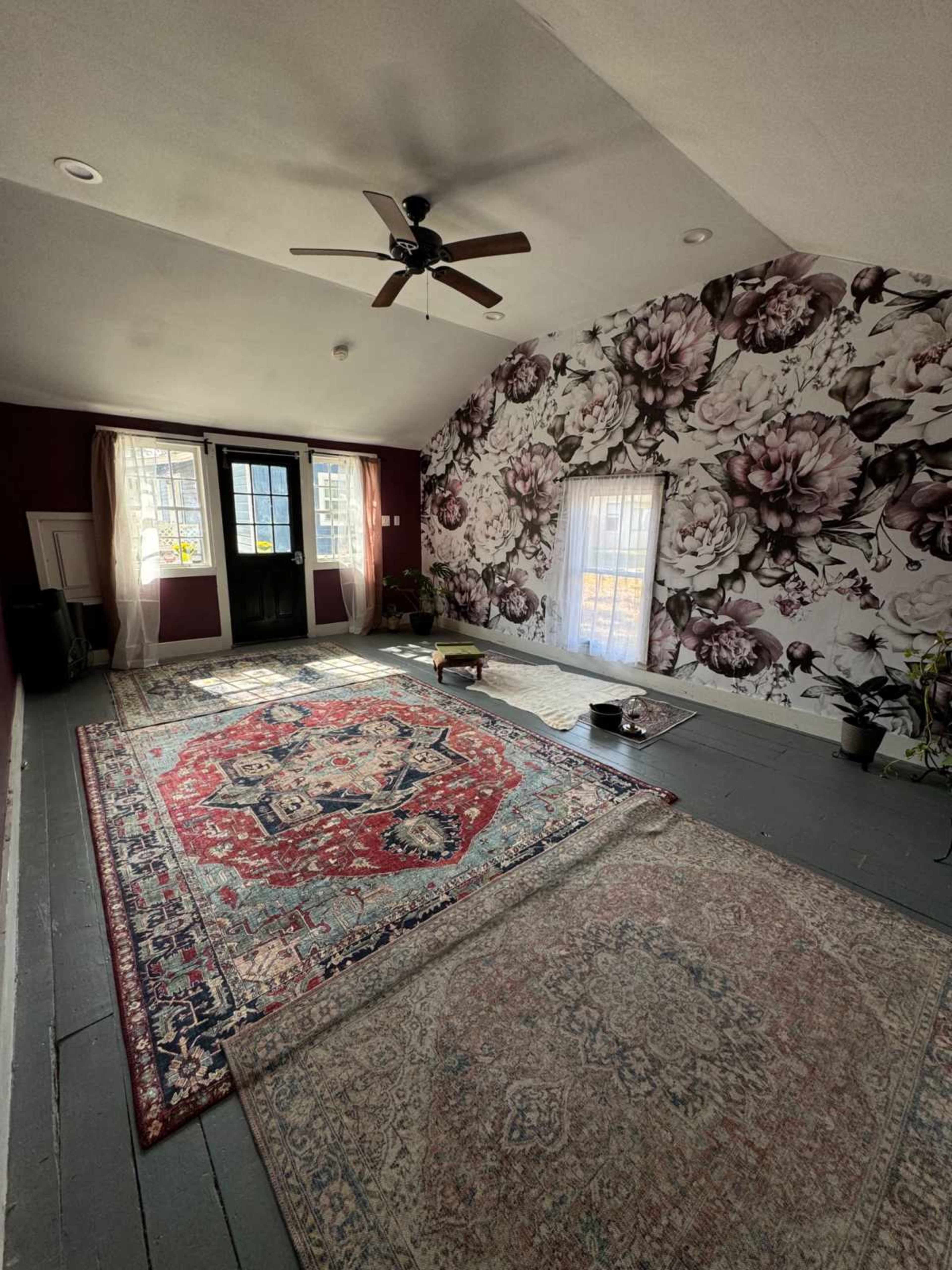 The image shows a spacious room with a floral wallpaper, hardwood floors, and several area rugs, along with a ceiling fan and two windows.