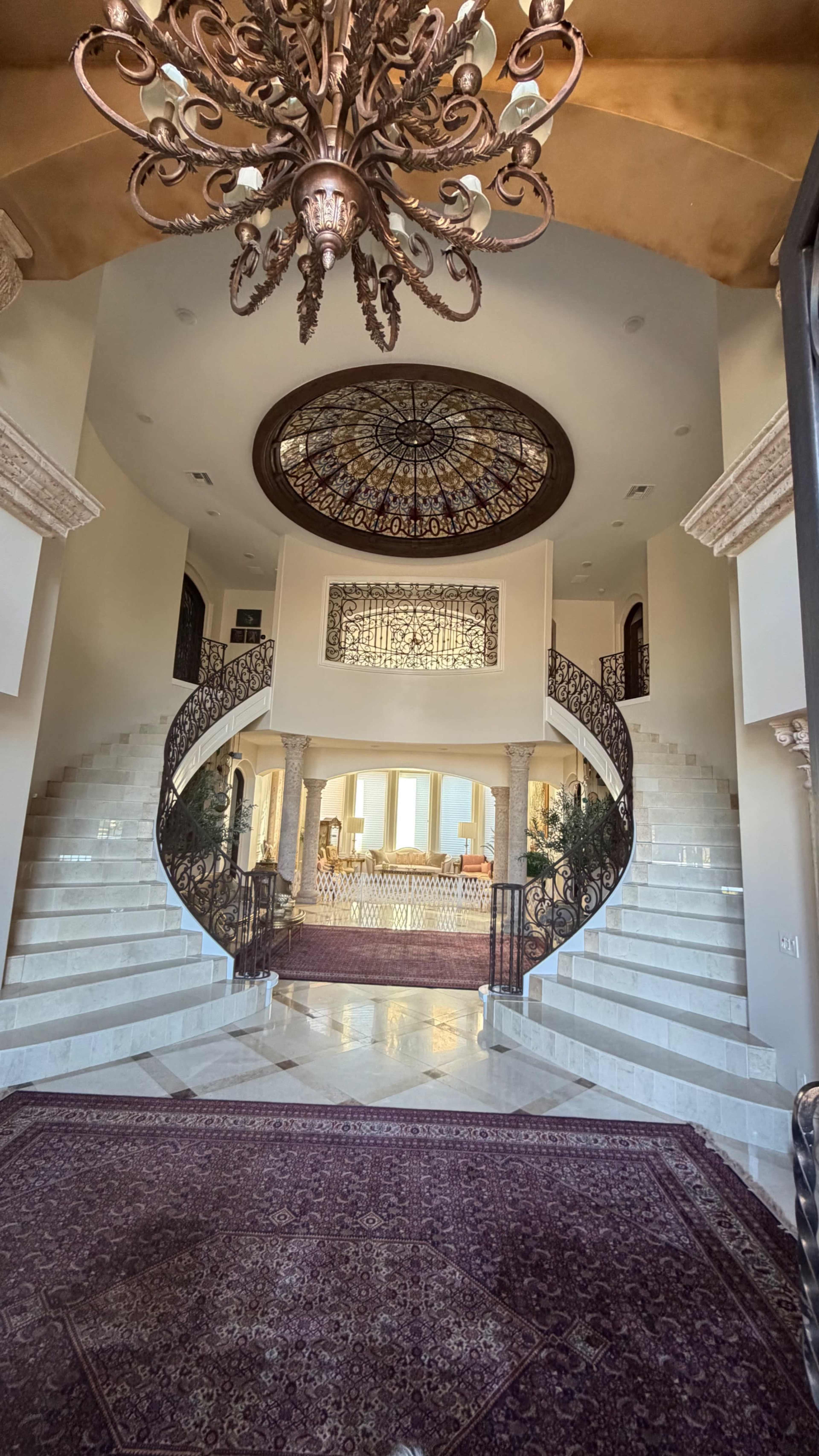 The image shows a grand foyer with a large chandelier, ornate staircase, and decorative ceiling.