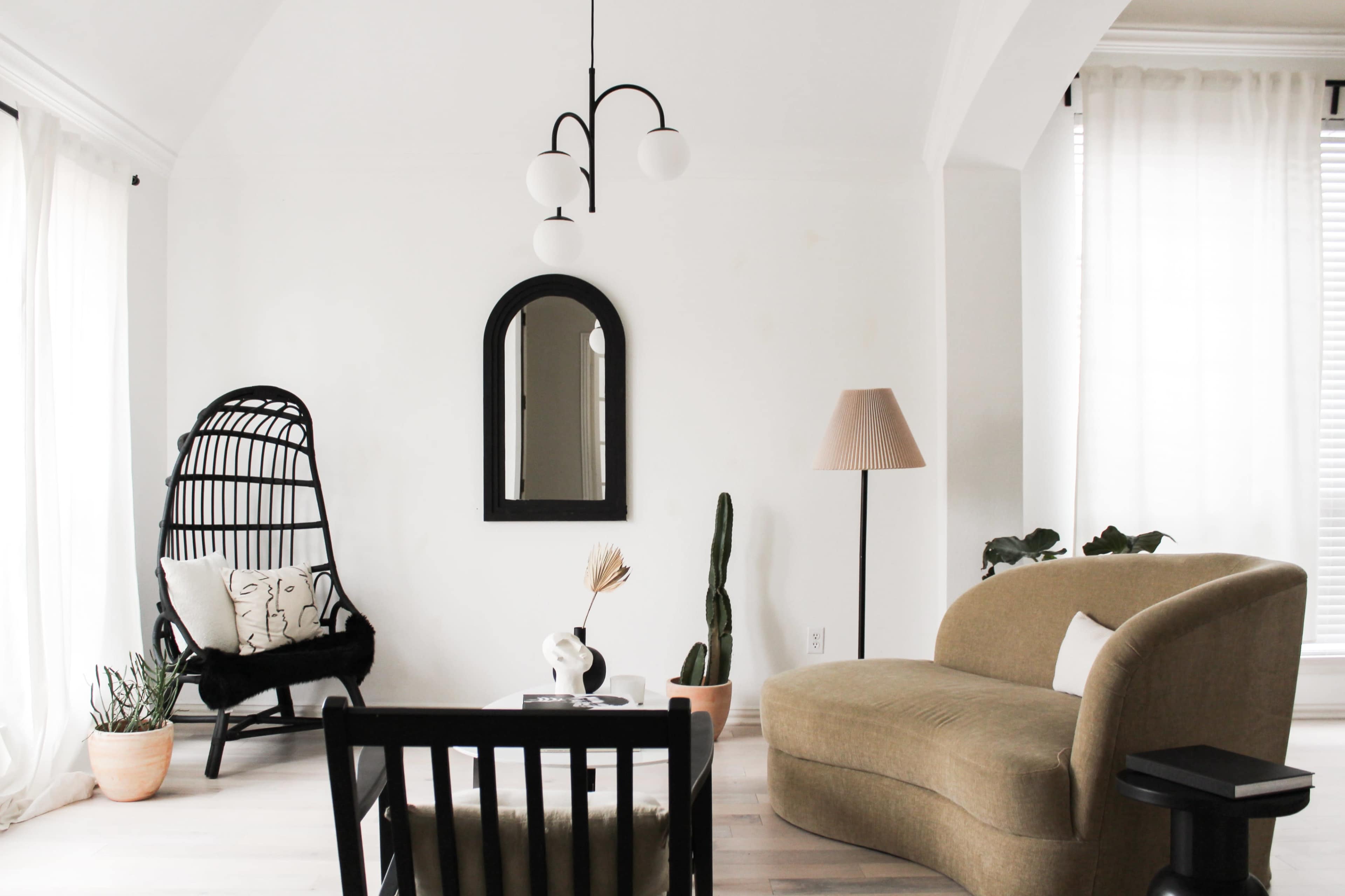 The image shows a modern living room featuring a curved sofa, a black chair, a lamp, a mirror, and various plants arranged against a white wall.
