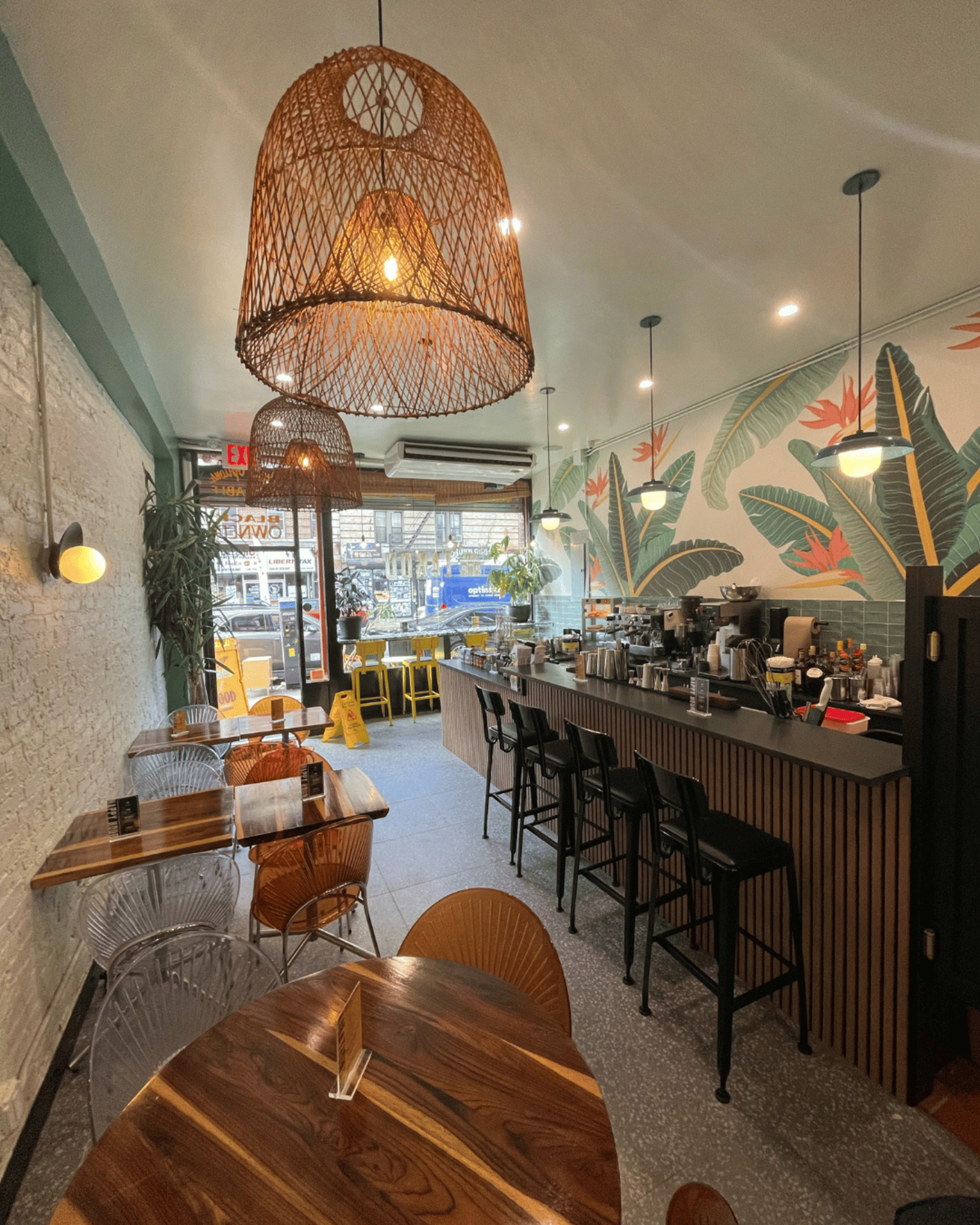 The image shows a cafe interior with wooden tables, a counter with bar stools, and a wall adorned with tropical-themed wallpaper.