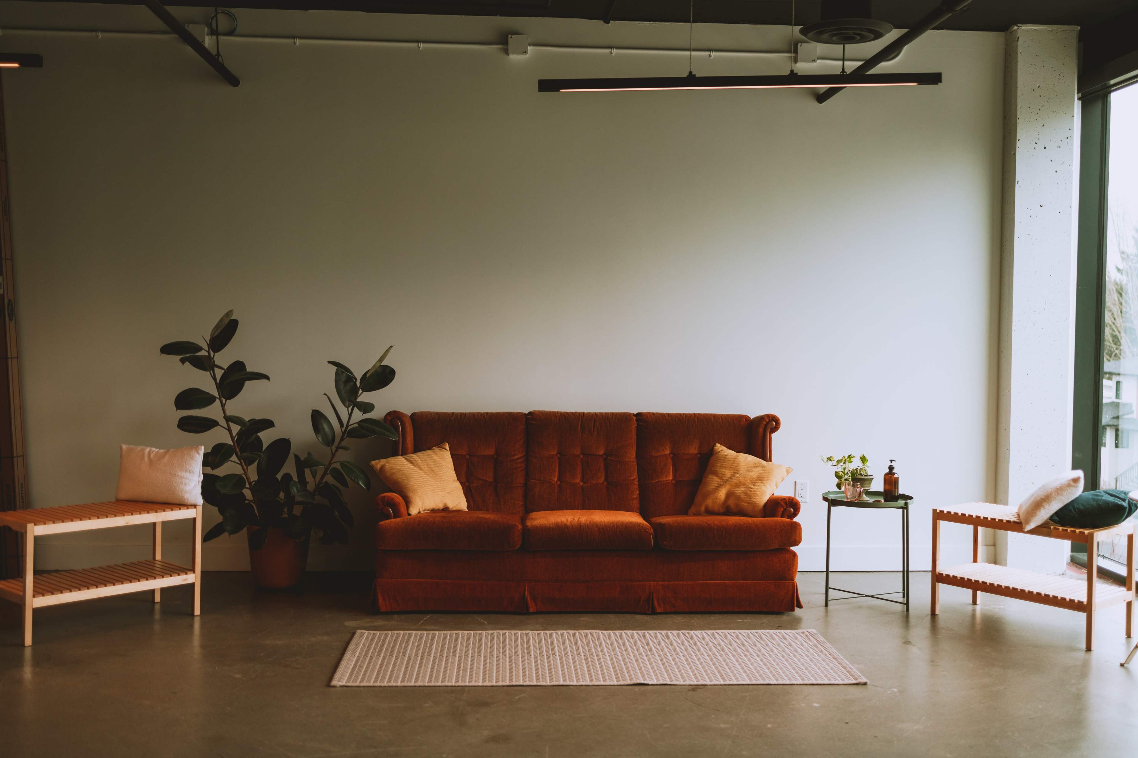 An orange sofa with two cushions, flanked by plants and wooden side tables in a spacious room with minimal decor.