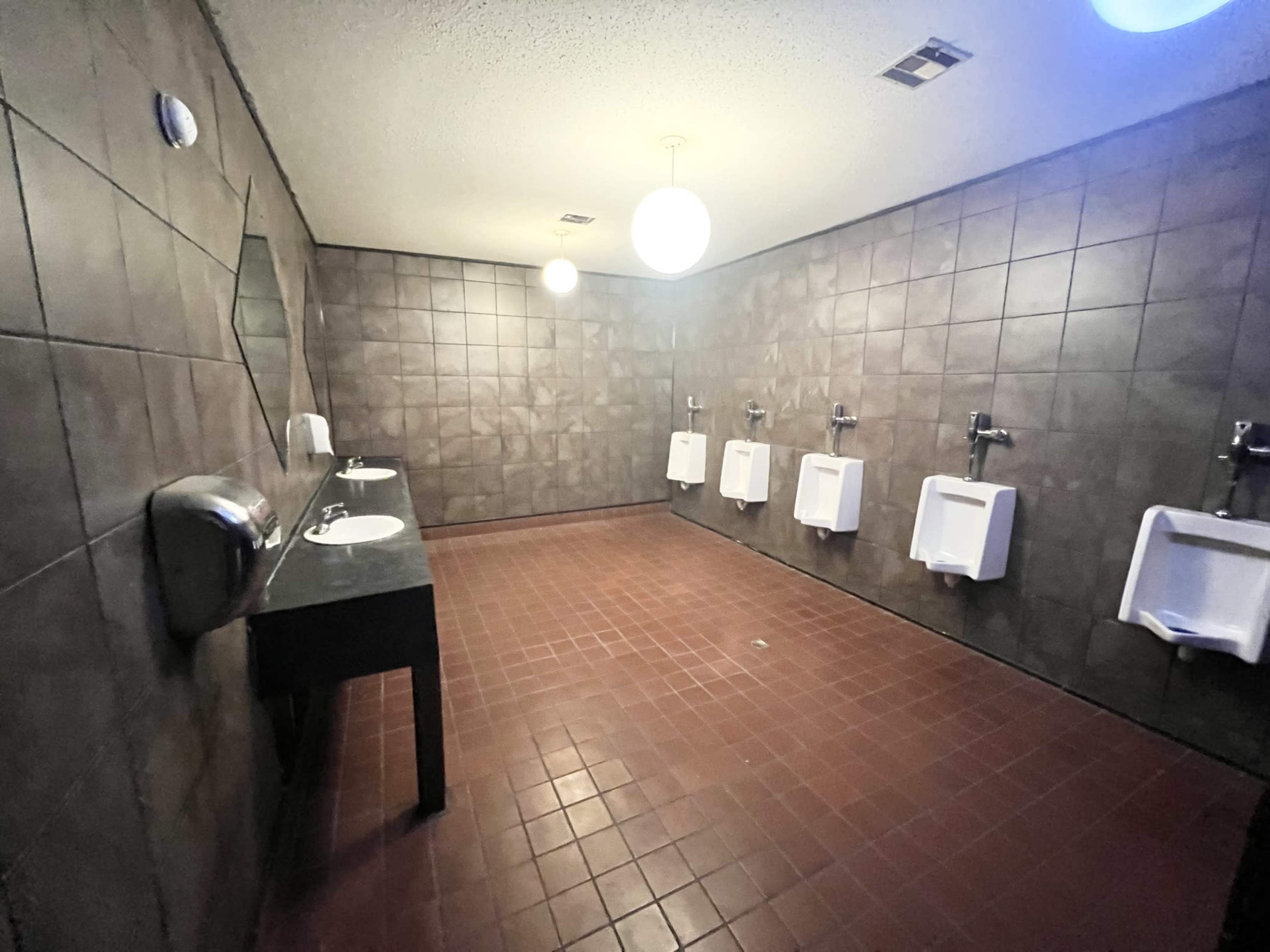 A men's restroom features three urinals mounted on the wall and two sinks with a mirror above them.
