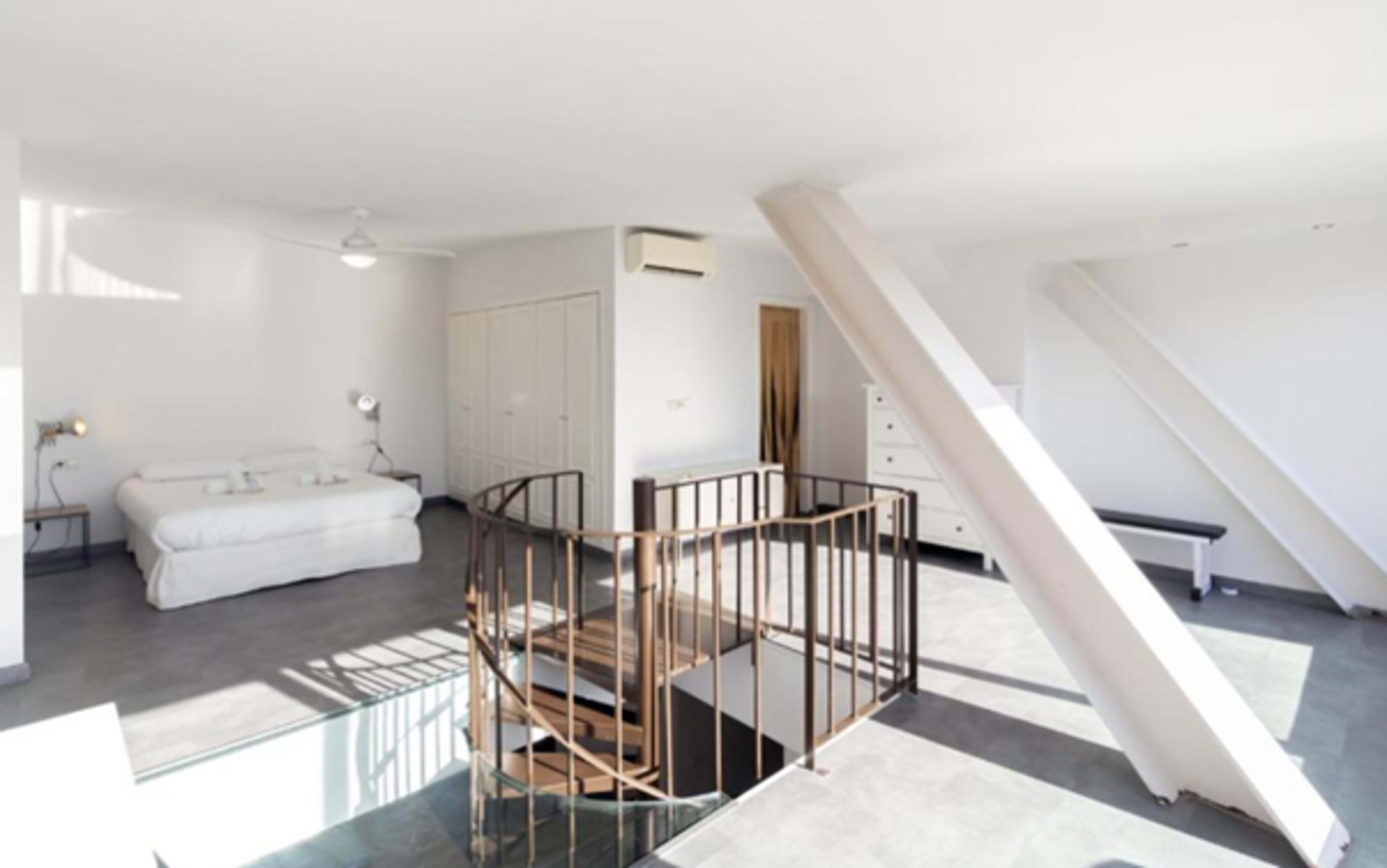 The image shows a modern, minimalist interior space with a white bed, a staircase, and various storage units.