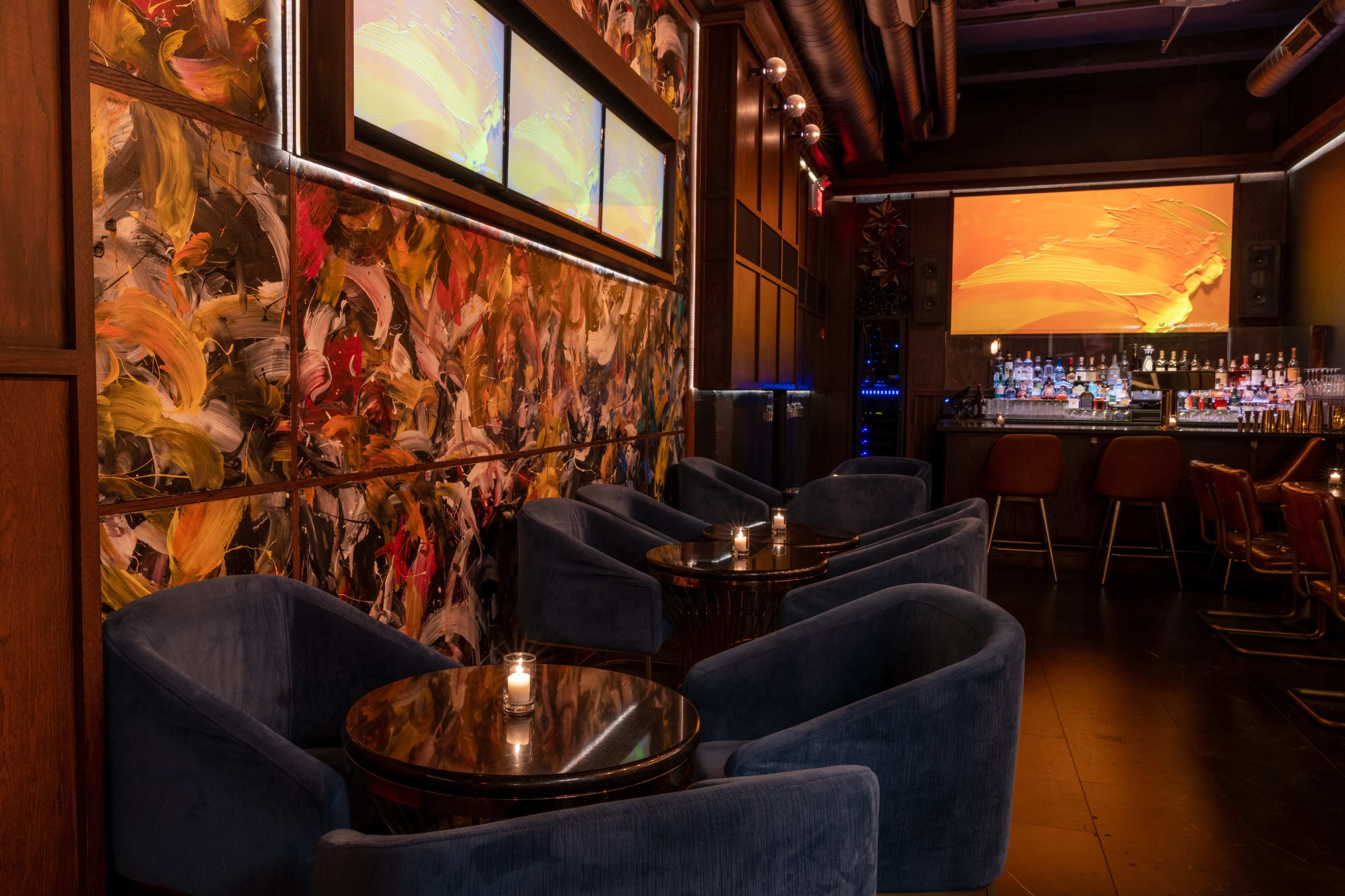 A modern lounge features plush blue seating arranged around small round tables, with a vibrant, abstract mural on the wall and a bar area in the background.