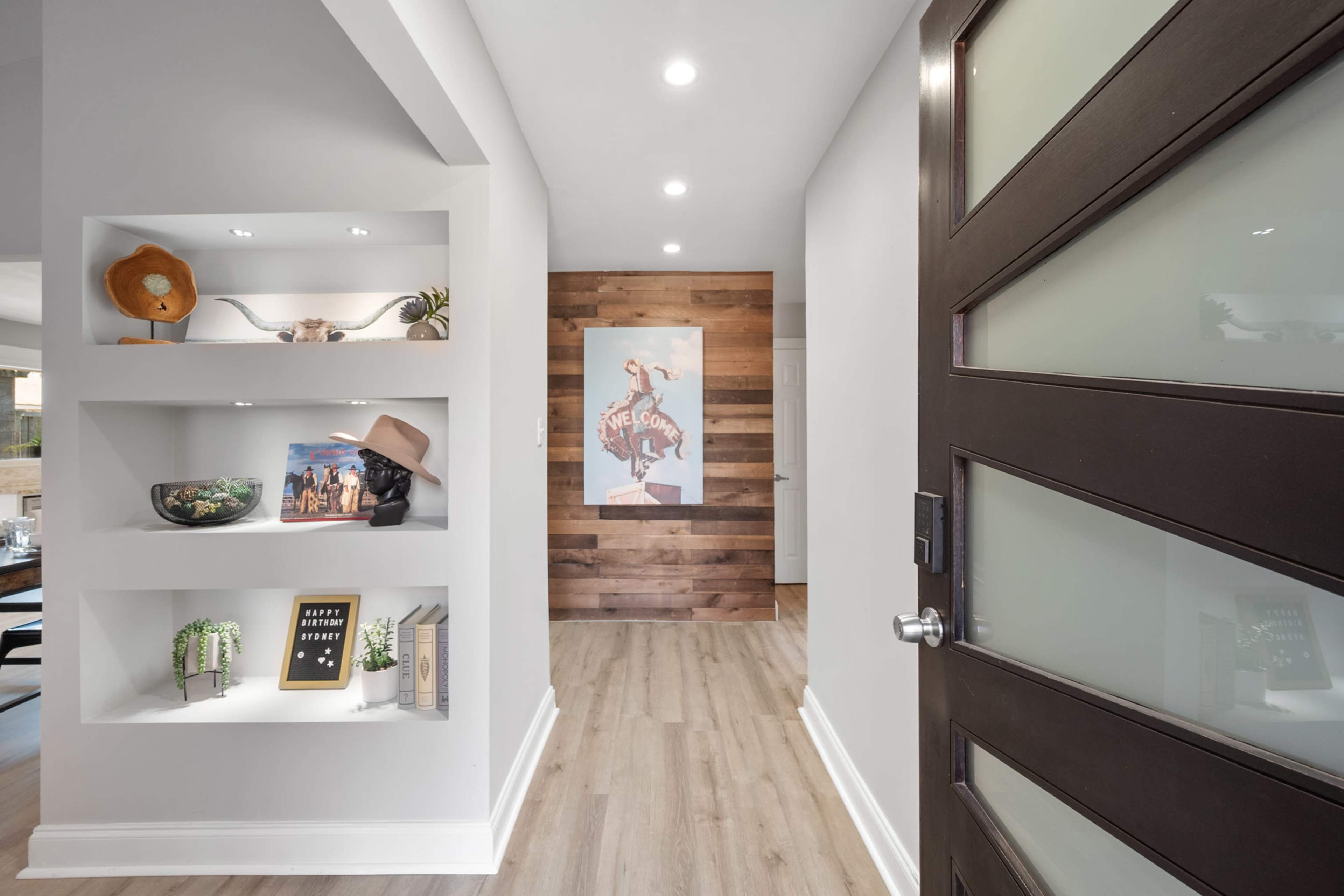 A hallway features a modern entry door, built-in shelves with decorative items, and a wood-paneled accent wall displaying artwork.