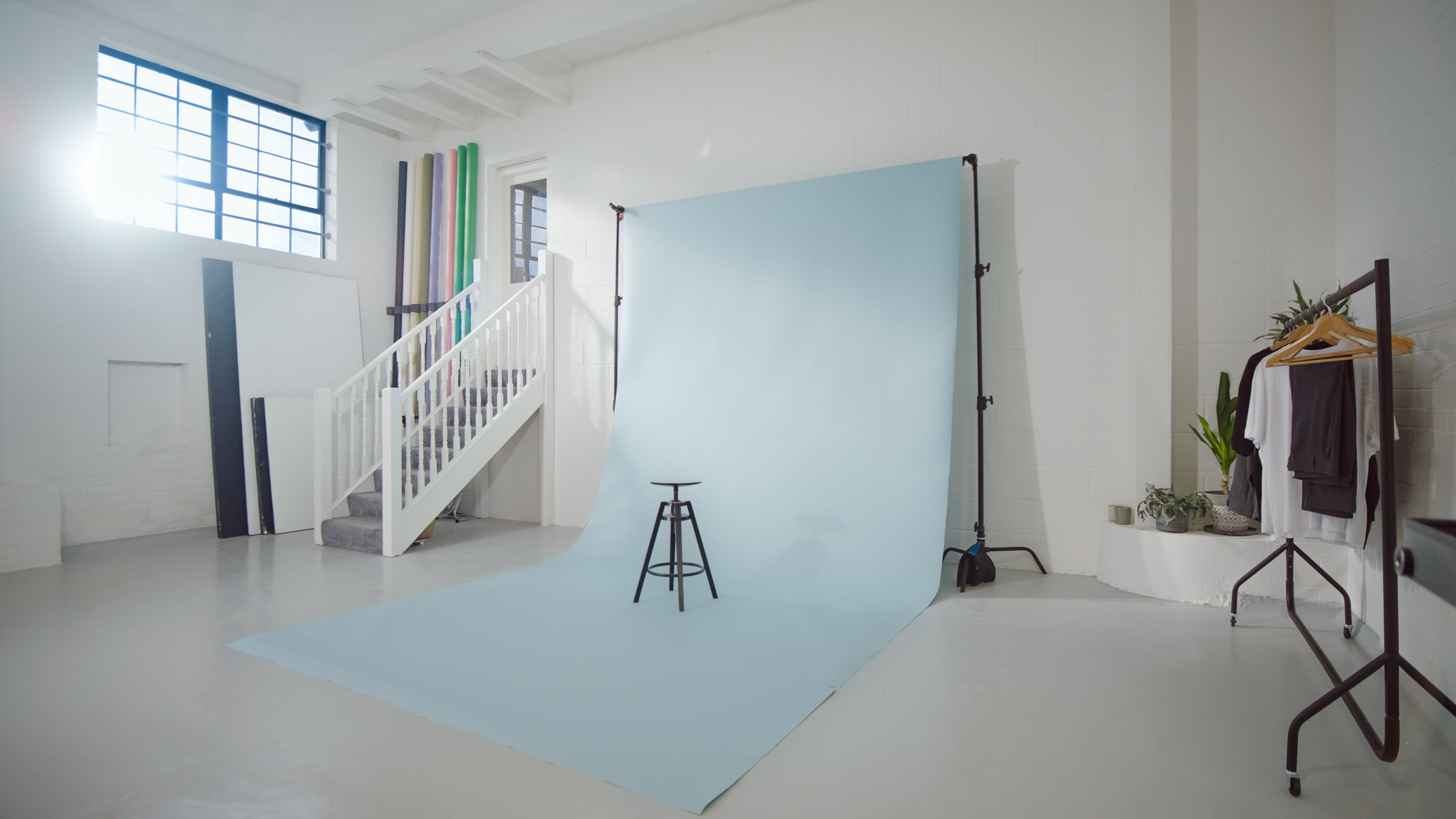 The image shows a photography studio with a light blue backdrop, a black stool positioned in front, and various colored backdrops neatly arranged in the background.