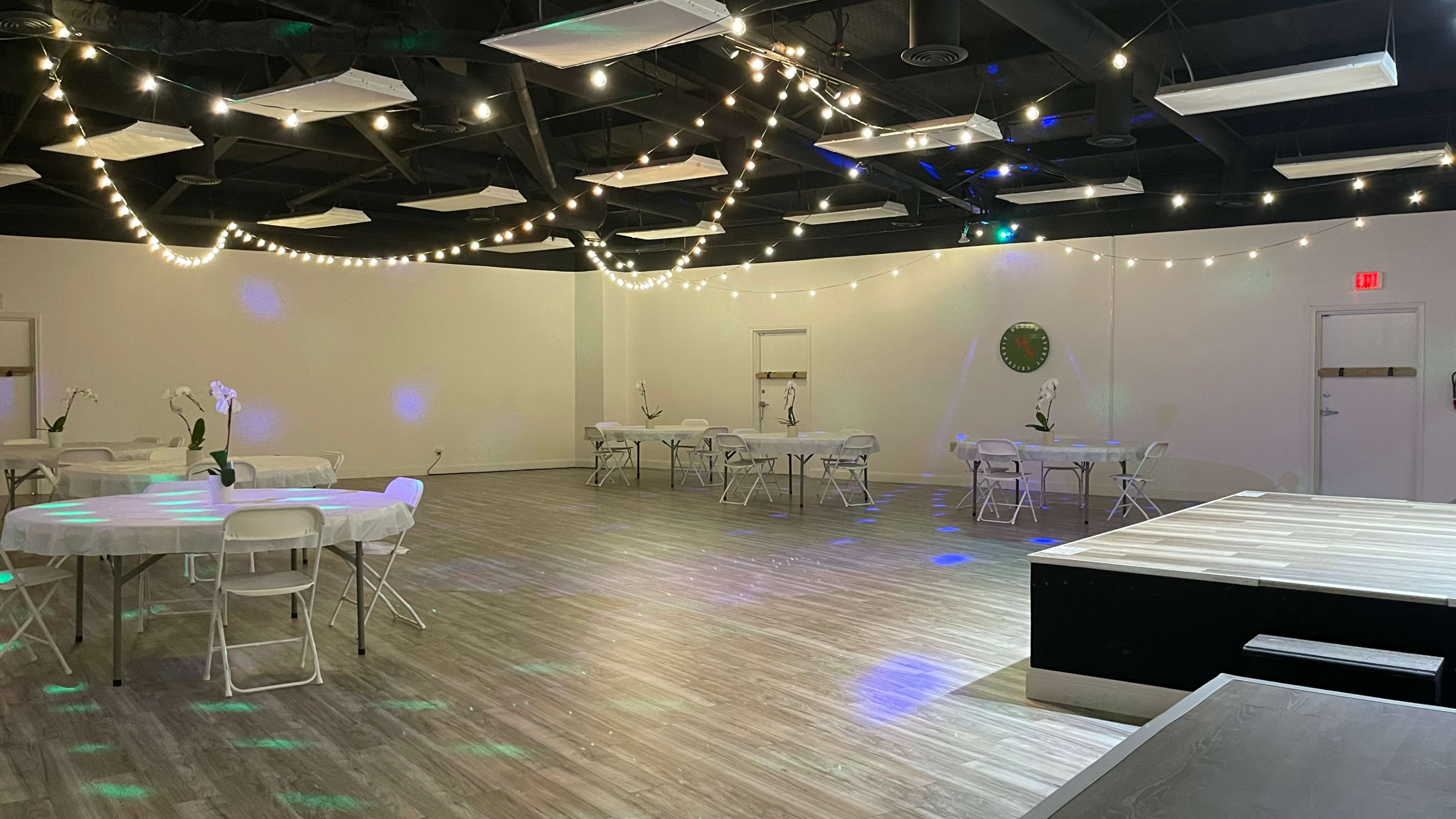 A spacious event room features round tables with chairs, ambient string lights, and a small stage area, all set against a plain white backdrop.
