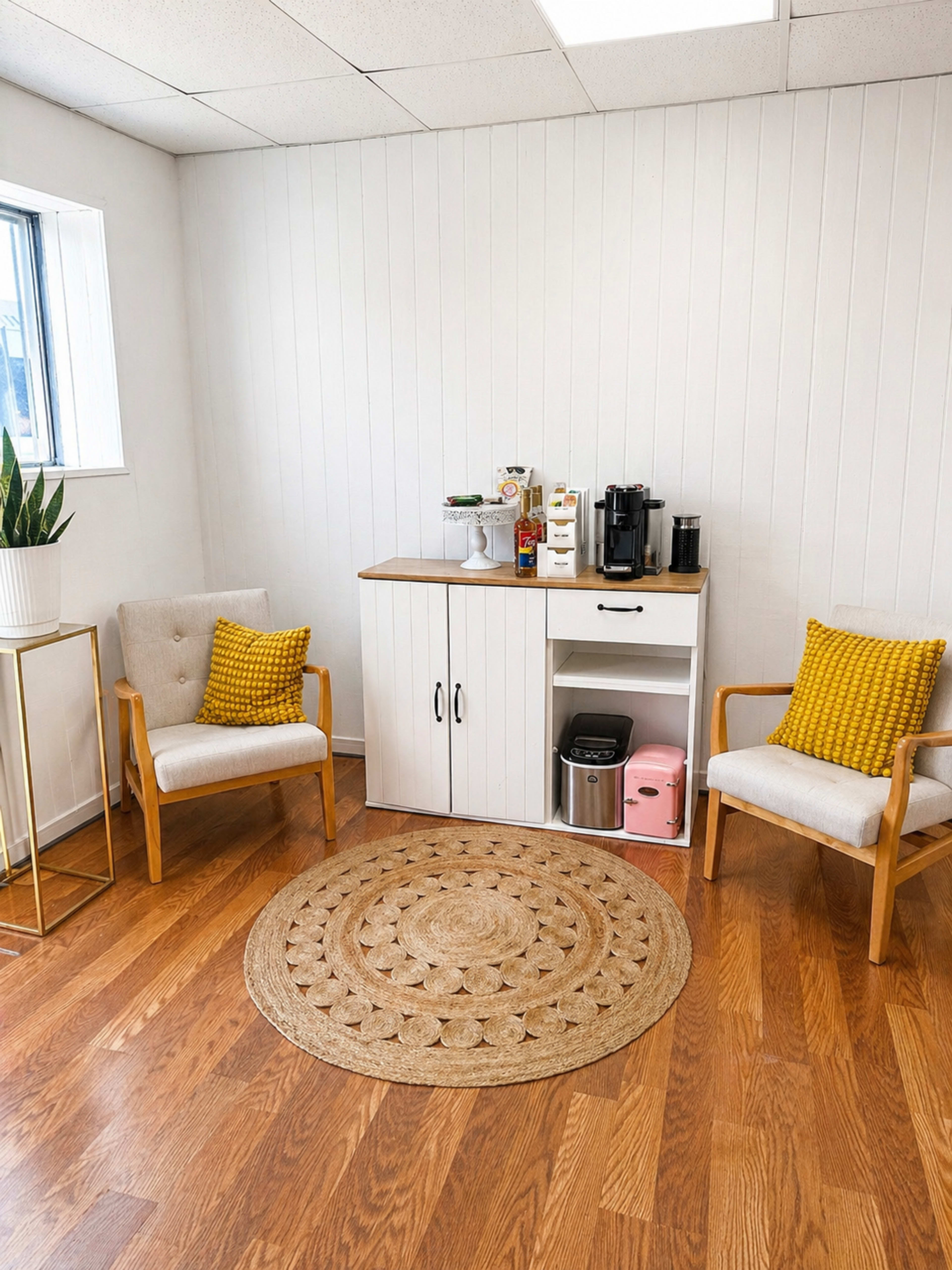 The image shows a small, well-lit corner of a room with two chairs, a round rug, and a cabinet containing a coffee maker and other supplies.