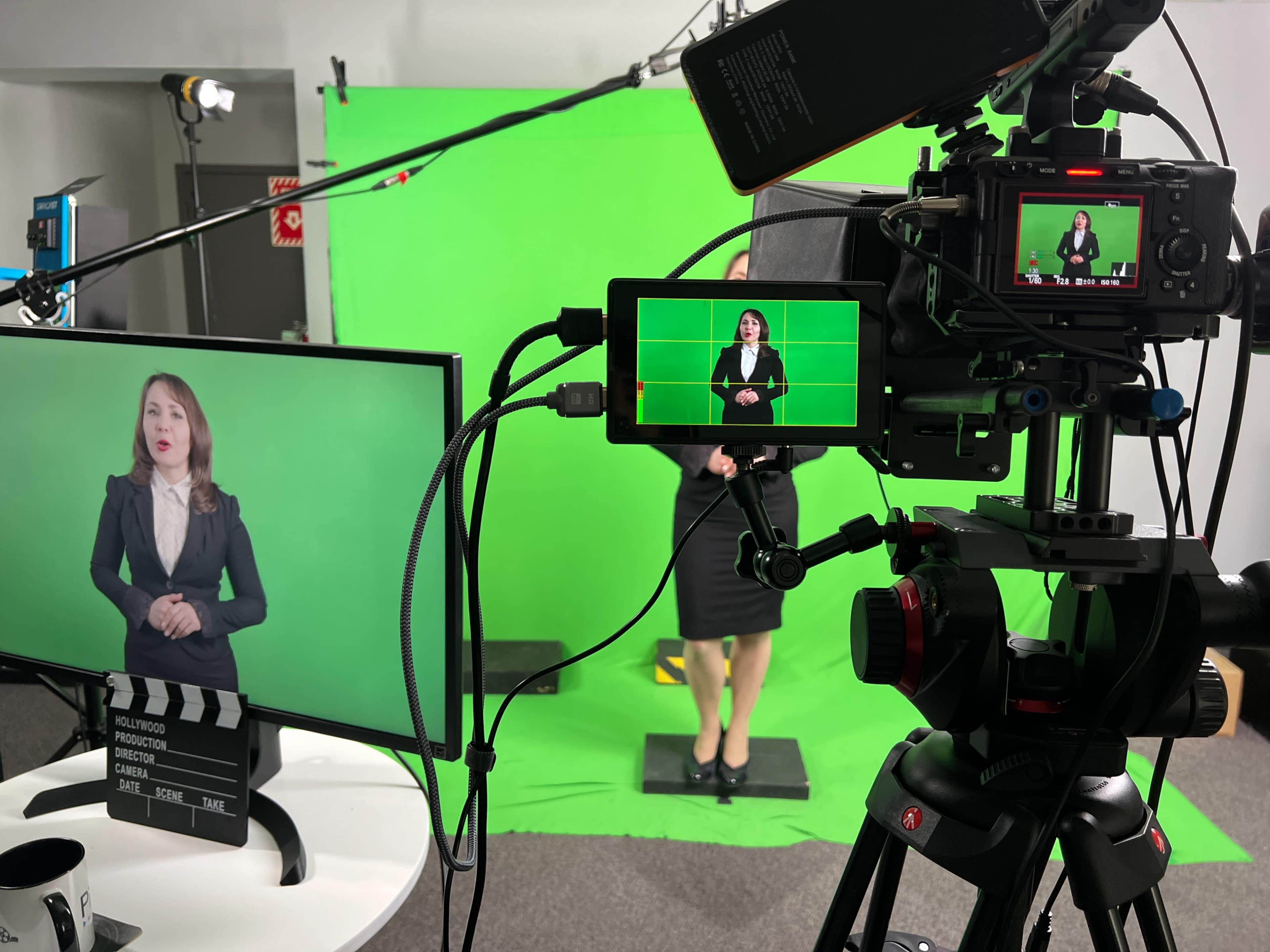 A woman in a black suit stands in front of a green screen while being recorded by cameras on tripods and a monitor displaying her image.