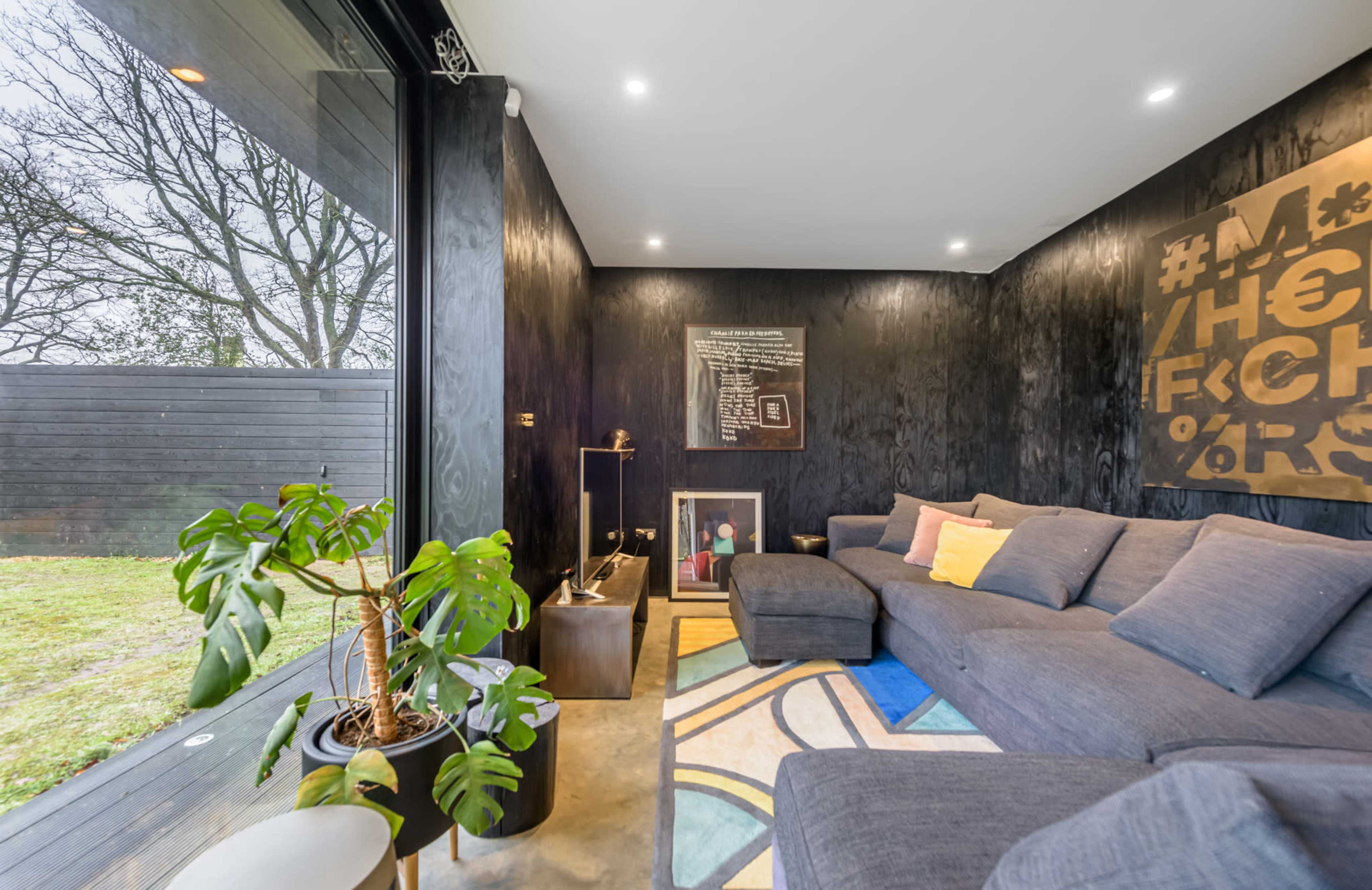 The room features a modern grey sofa, colorful rug, and large windows overlooking an outdoor space with trees.