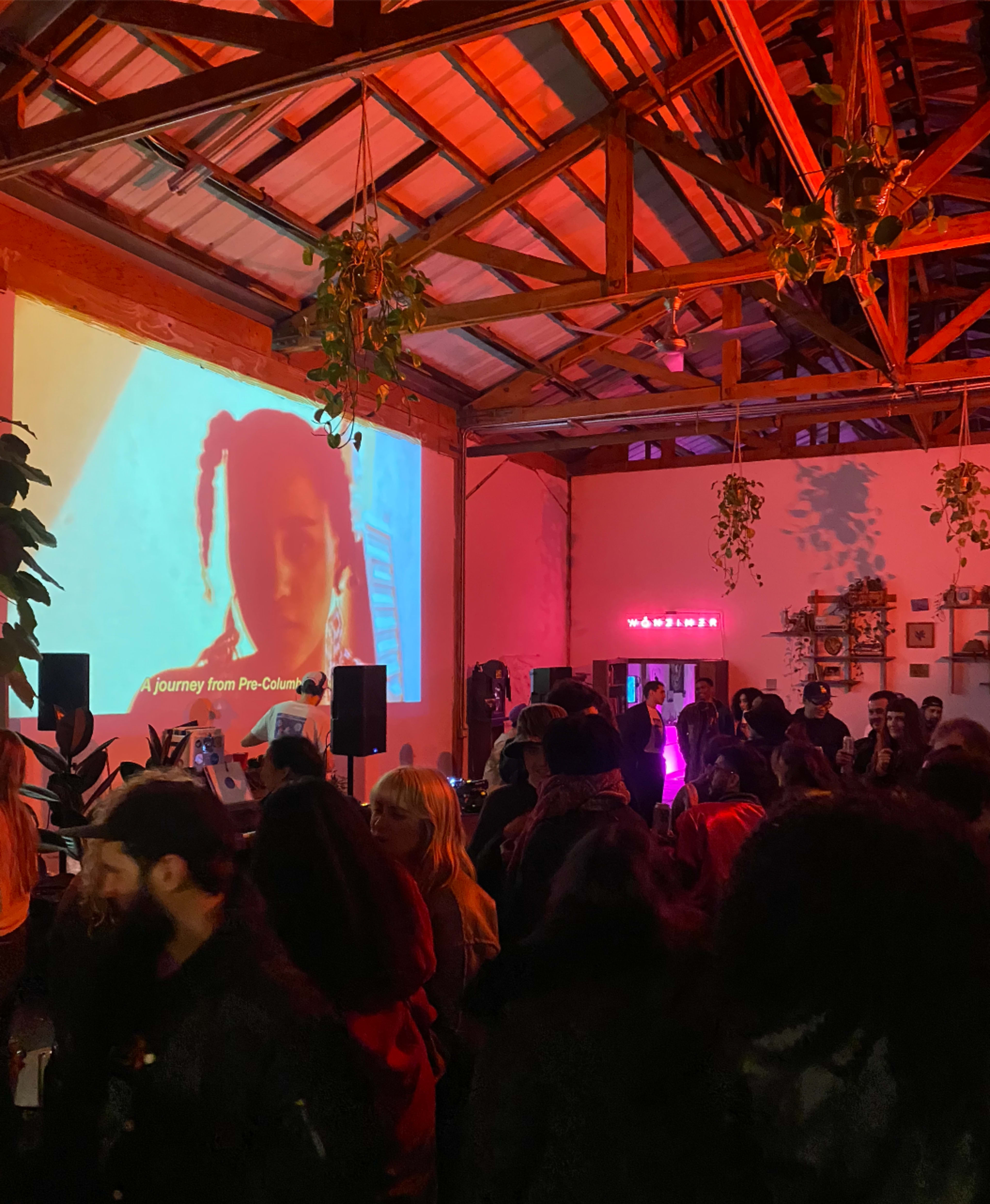 A crowd gathers in a warmly lit venue featuring a large screen displaying a video of a person, surrounded by plants and artistic decor.
