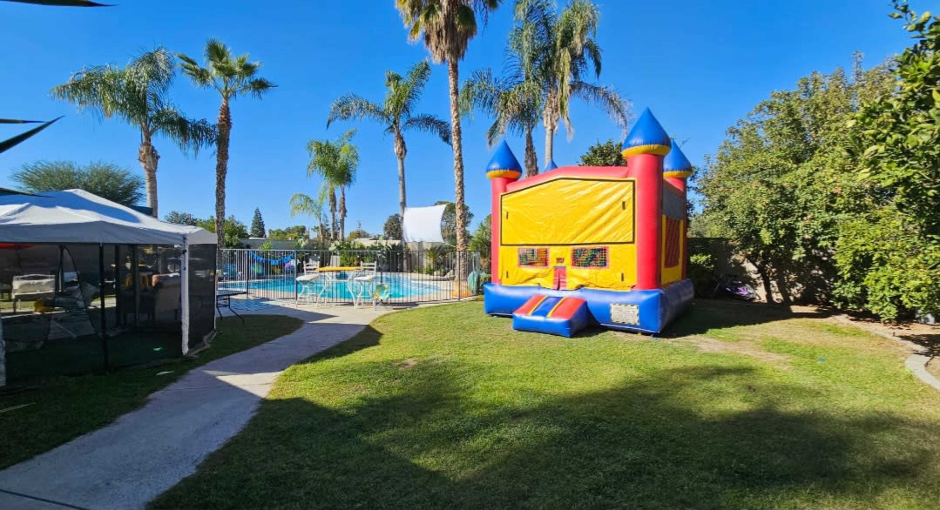 A brightly colored bounce house is set up on a lawn beside a pool, surrounded by palm trees and greenery.