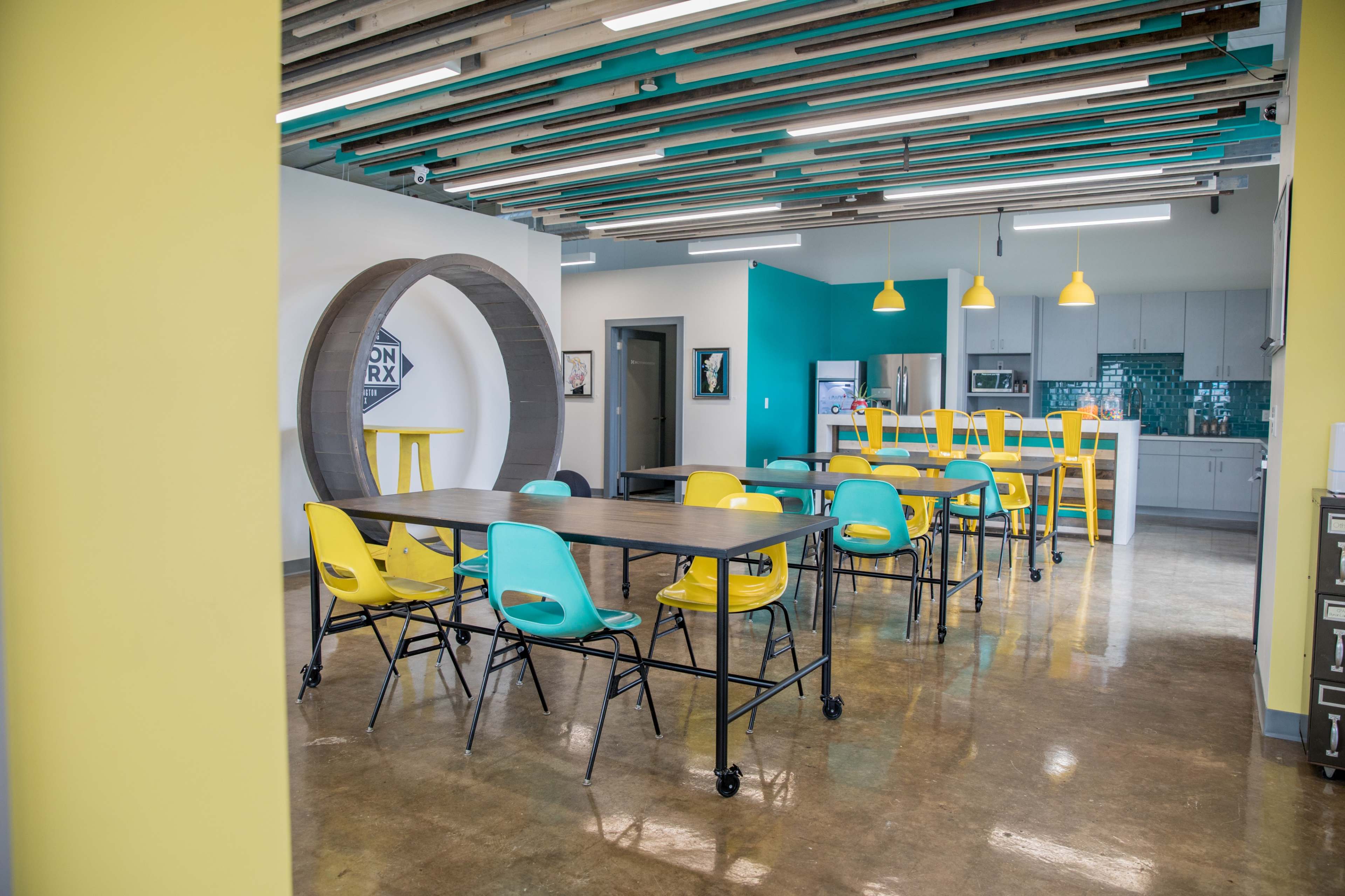 A modern office kitchen area featuring a dining space with colorful chairs and a large wooden table, along with kitchen appliances and bright wall accents.