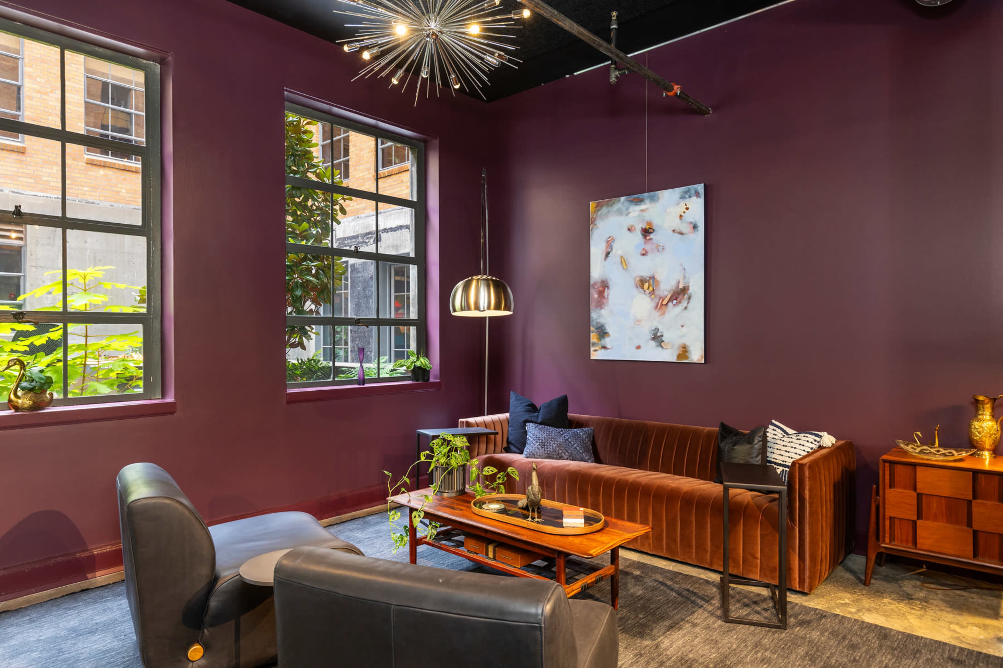 A modern lounge area features a velvet sofa, armchairs, a coffee table, and large windows that allow natural light to enter.