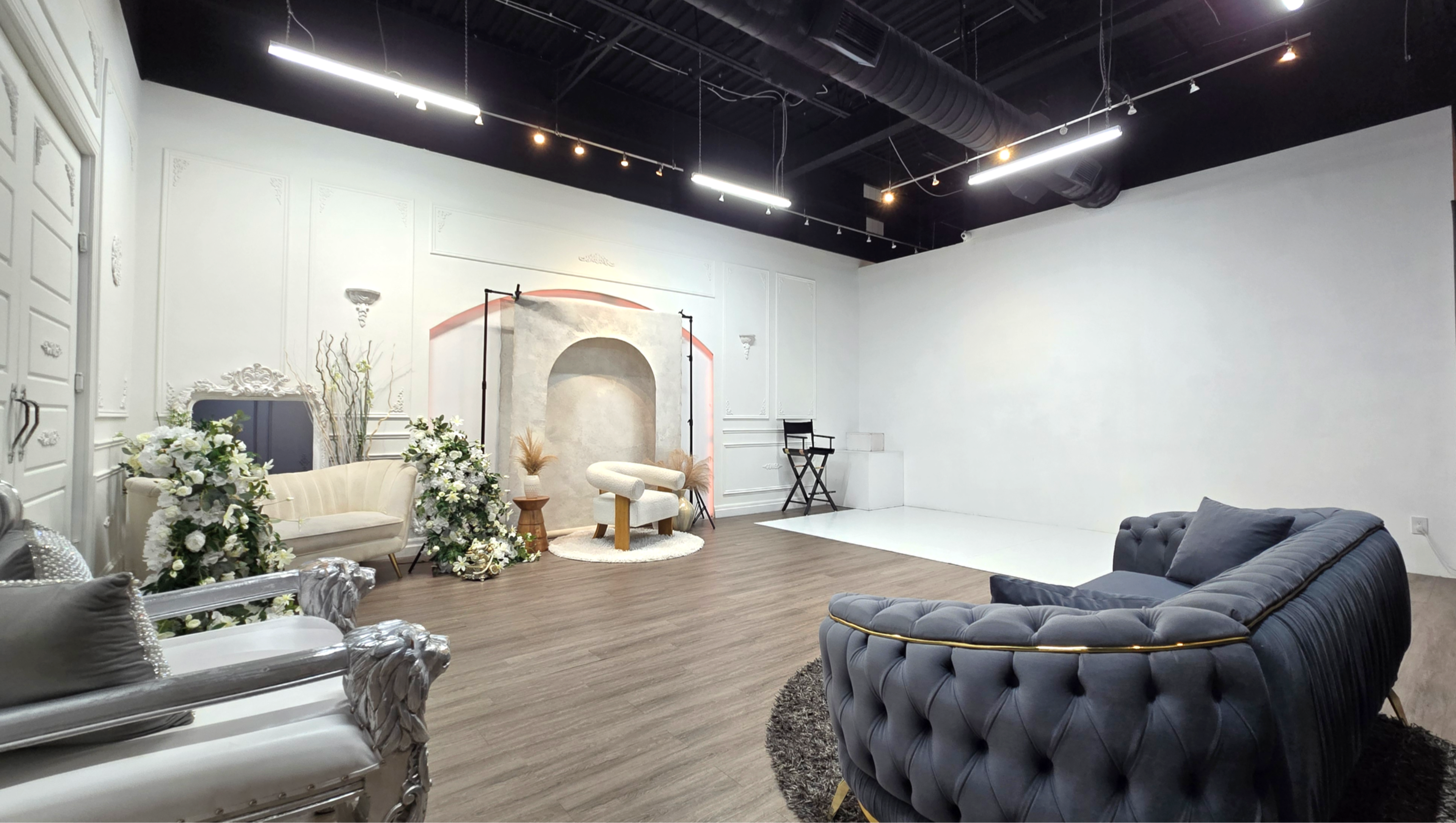 The image shows a spacious studio interior featuring a seating area with elegant furniture, a backdrop with a sculpted arch, and floral arrangements.