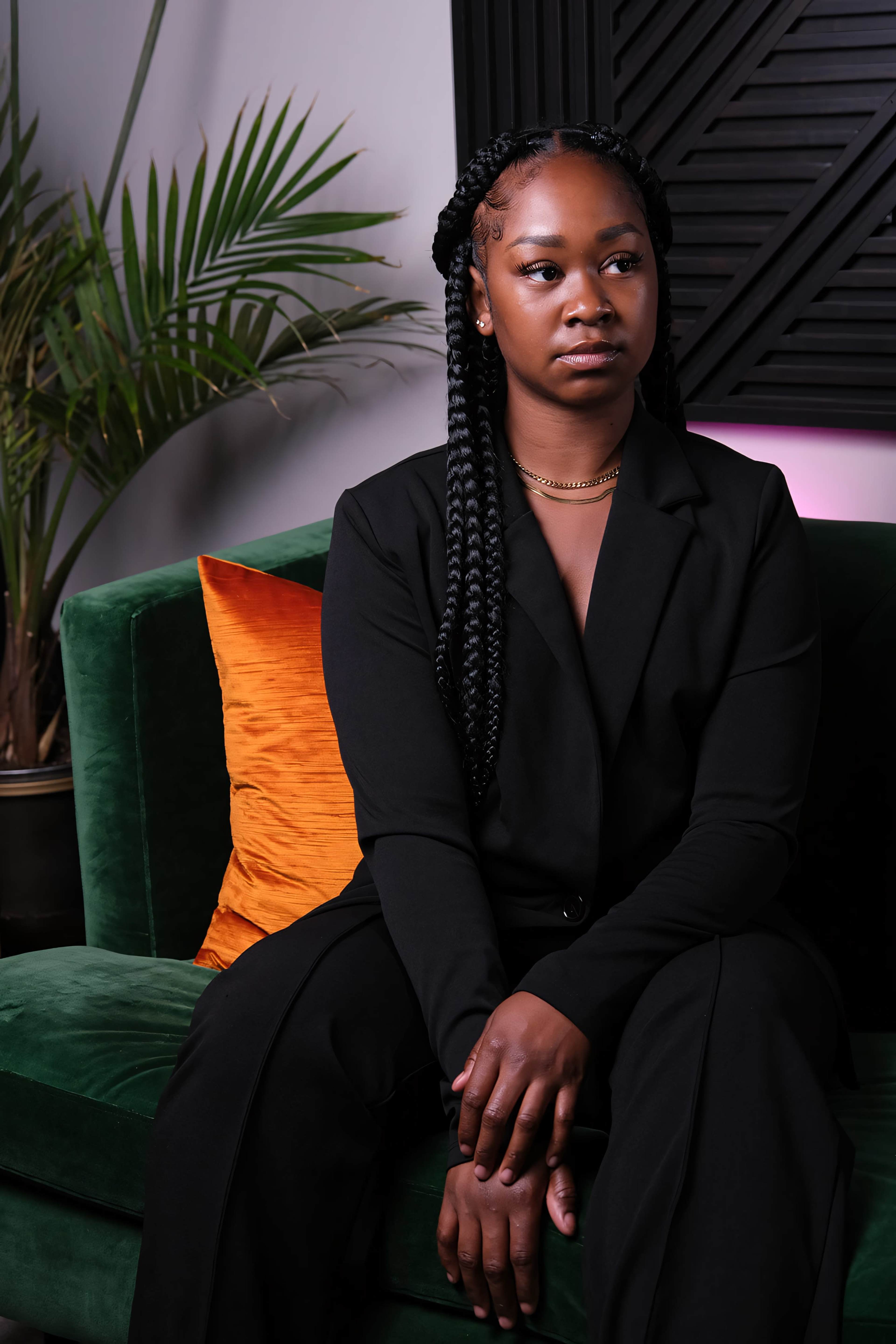 A woman with braided hair sits on a green velvet sofa next to an orange pillow, against a backdrop of a decorative wall and a potted plant.