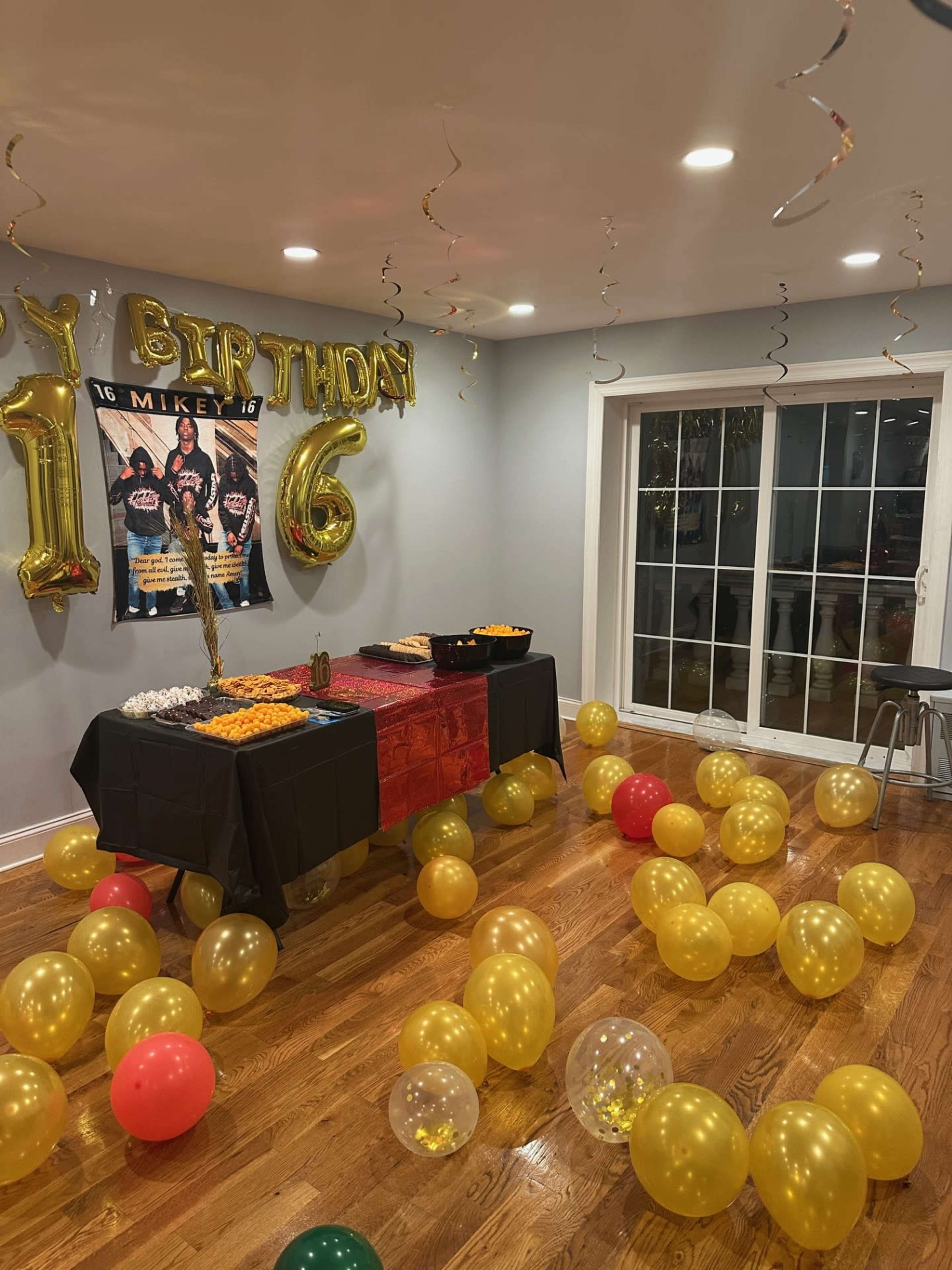 The image shows a birthday party setup with a black table covered in food, gold and red balloons scattered on the floor, and decorations overhead including the words "Happy Birthday" and a poster.