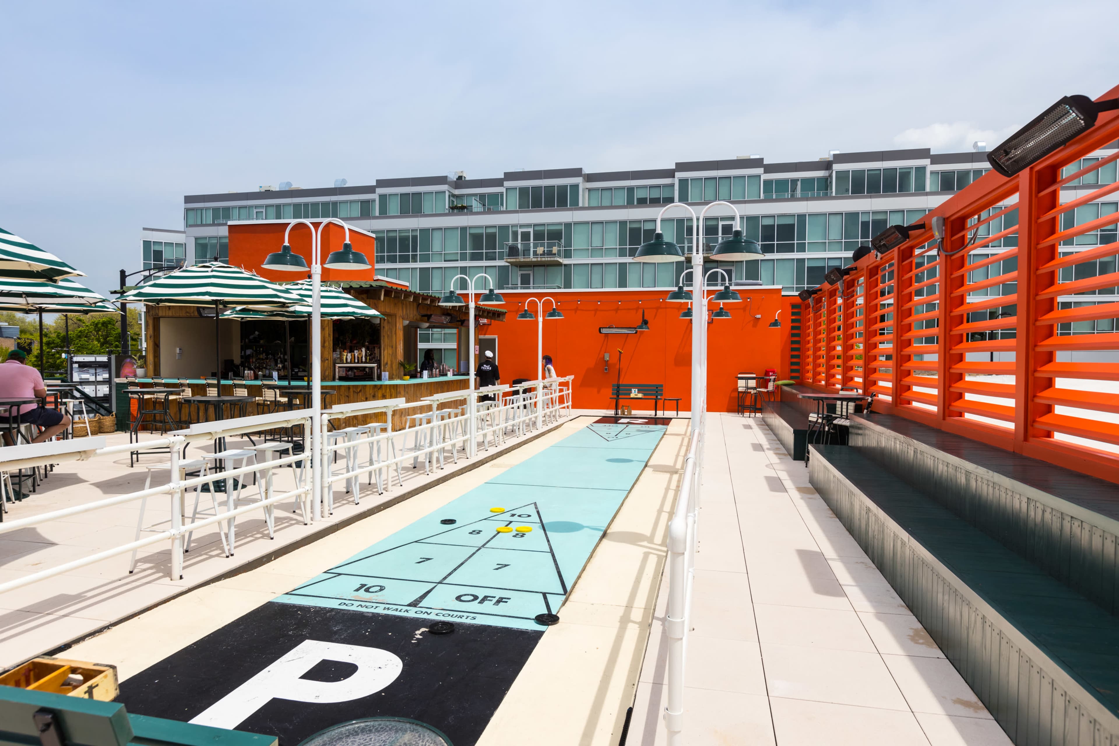The image shows a shuffleboard court with an adjacent bar area in a brightly colored outdoor space.