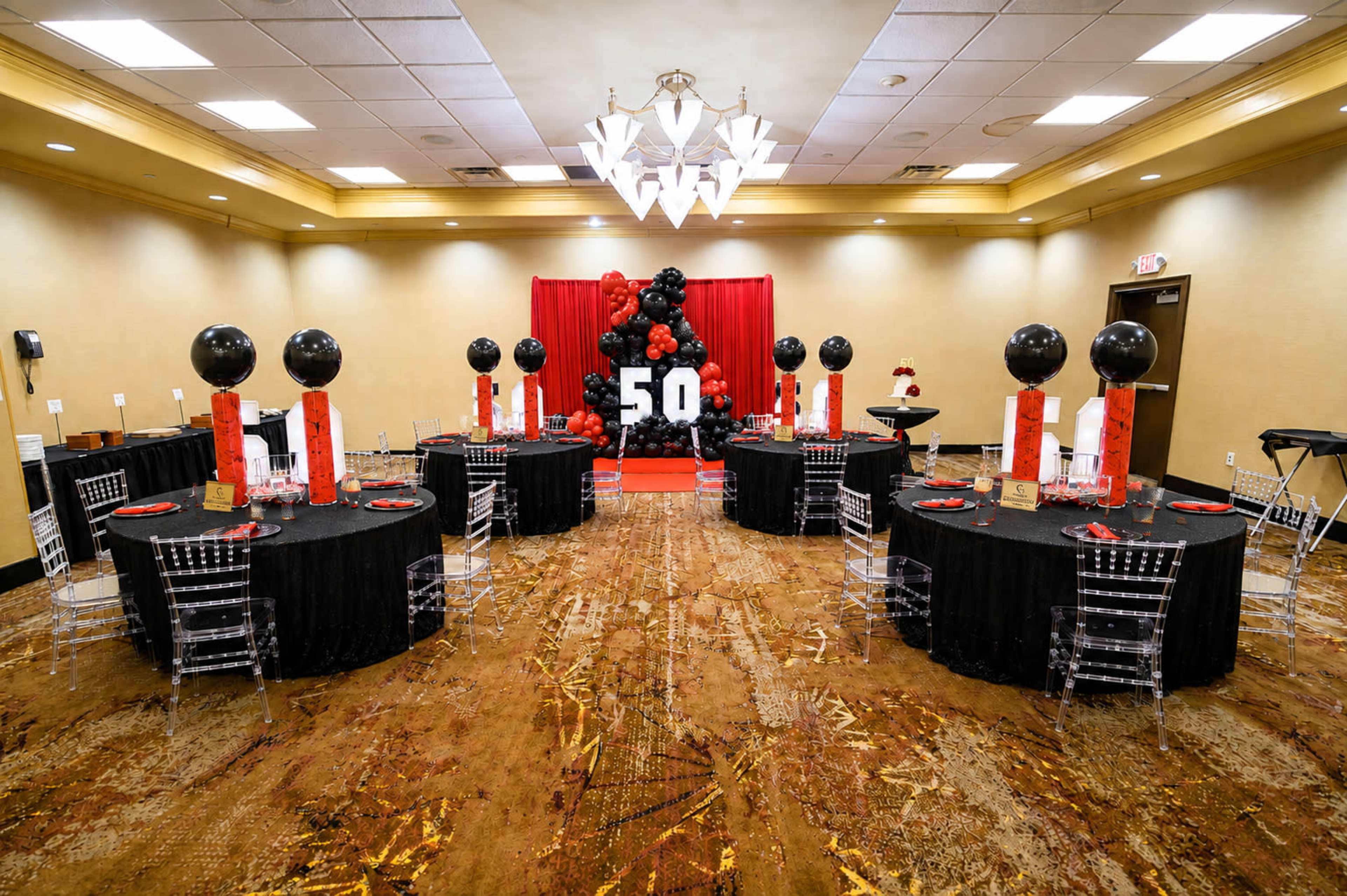 The image shows a banquet room arranged for a celebration with two rows of black tables, each decorated with red accents and topped with black balloons, set against a backdrop featuring the number "50."
