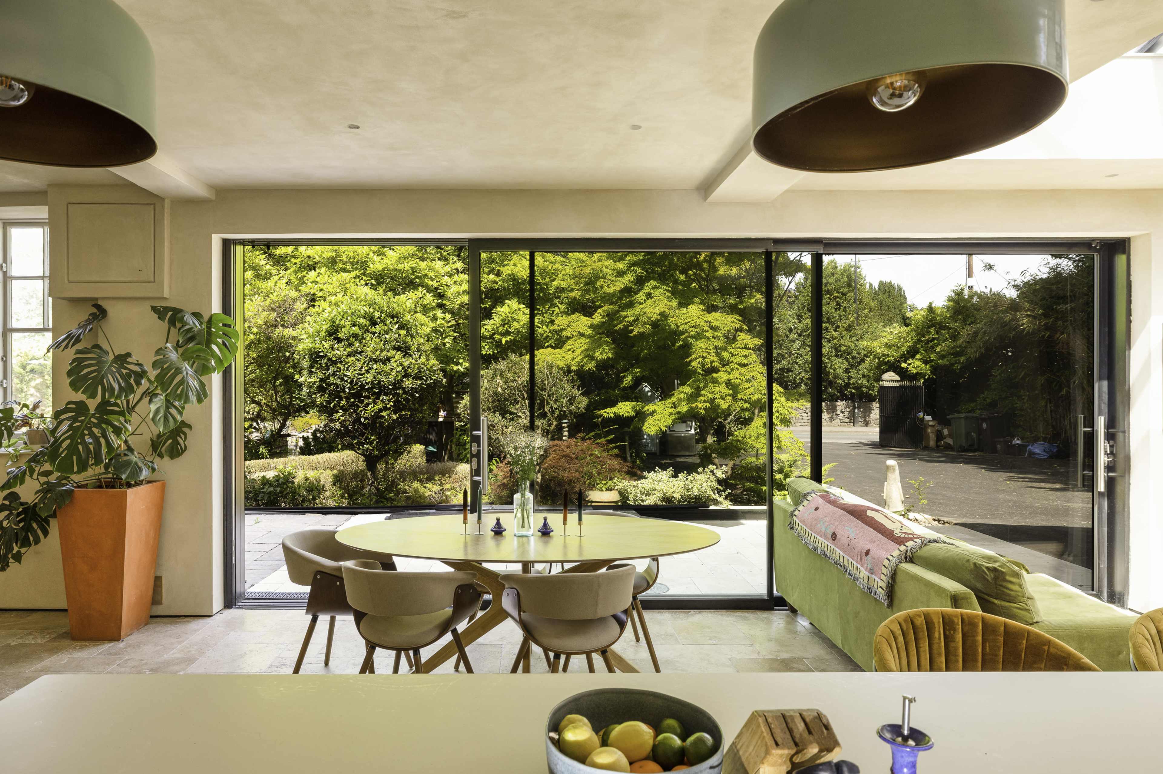 A modern kitchen area features a round dining table with chairs, surrounded by large windows that showcase a lush garden outside.