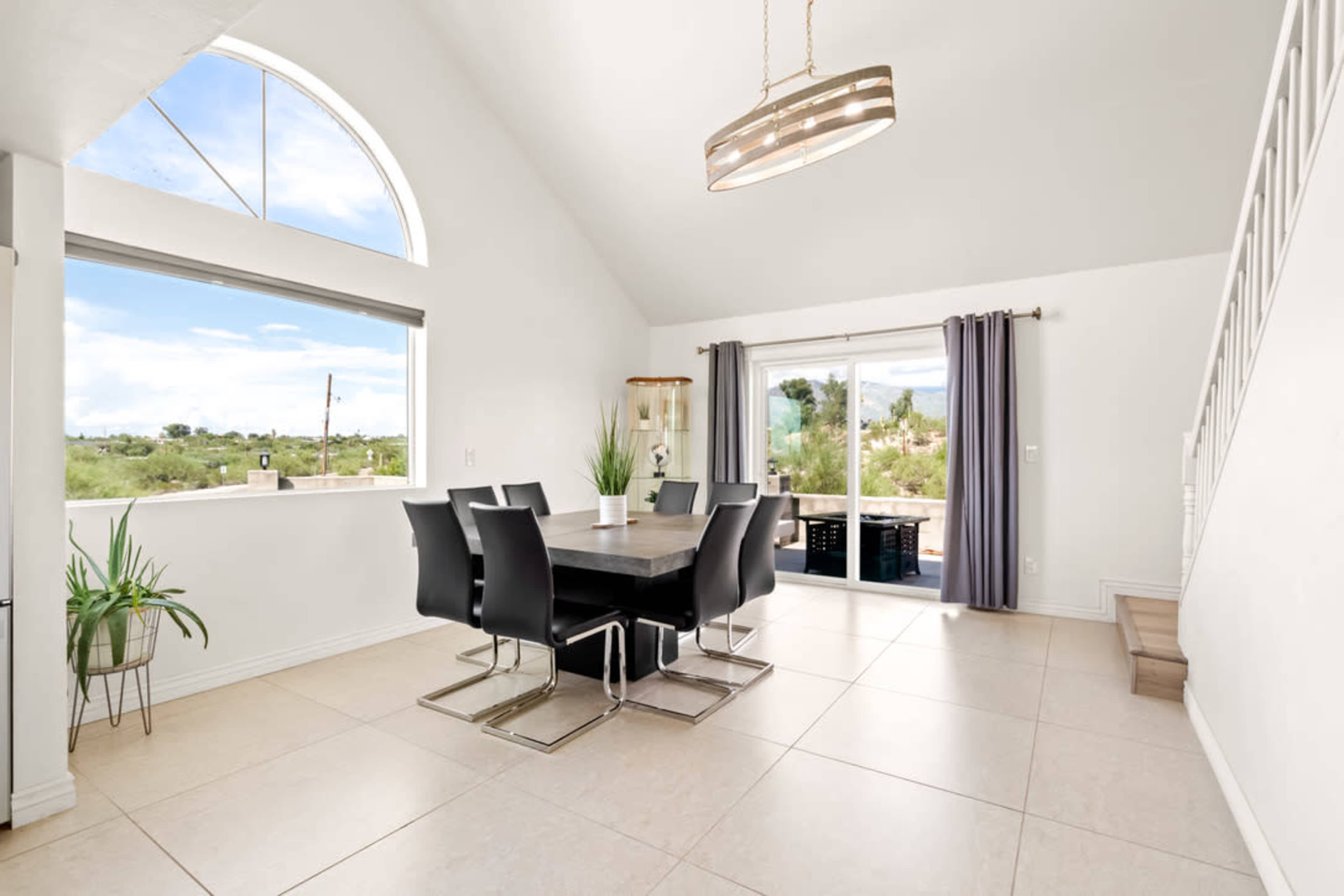 A dining room features a large table surrounded by modern chairs, with large windows showcasing an outdoor view.