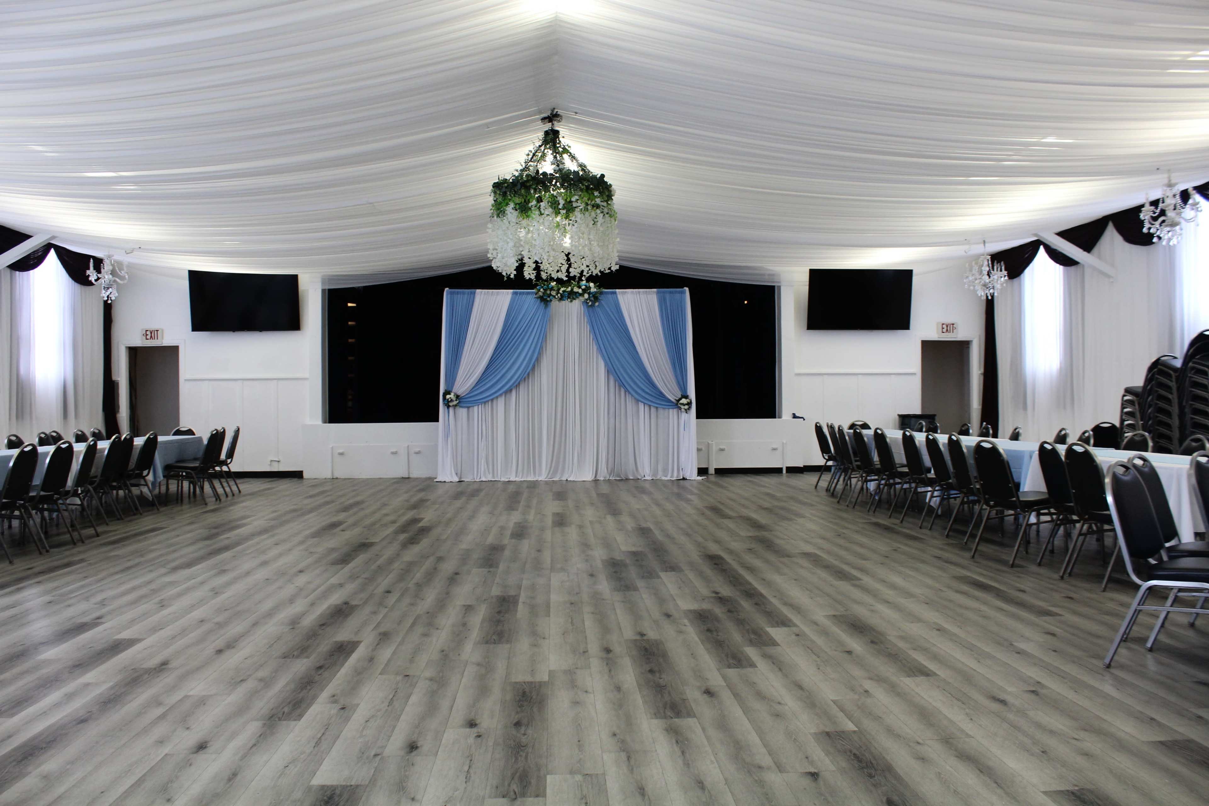 The image shows a spacious banquet hall with a high ceiling adorned with draped fabric, a decorative chandelier, and rows of tables and chairs arranged around a central stage area.