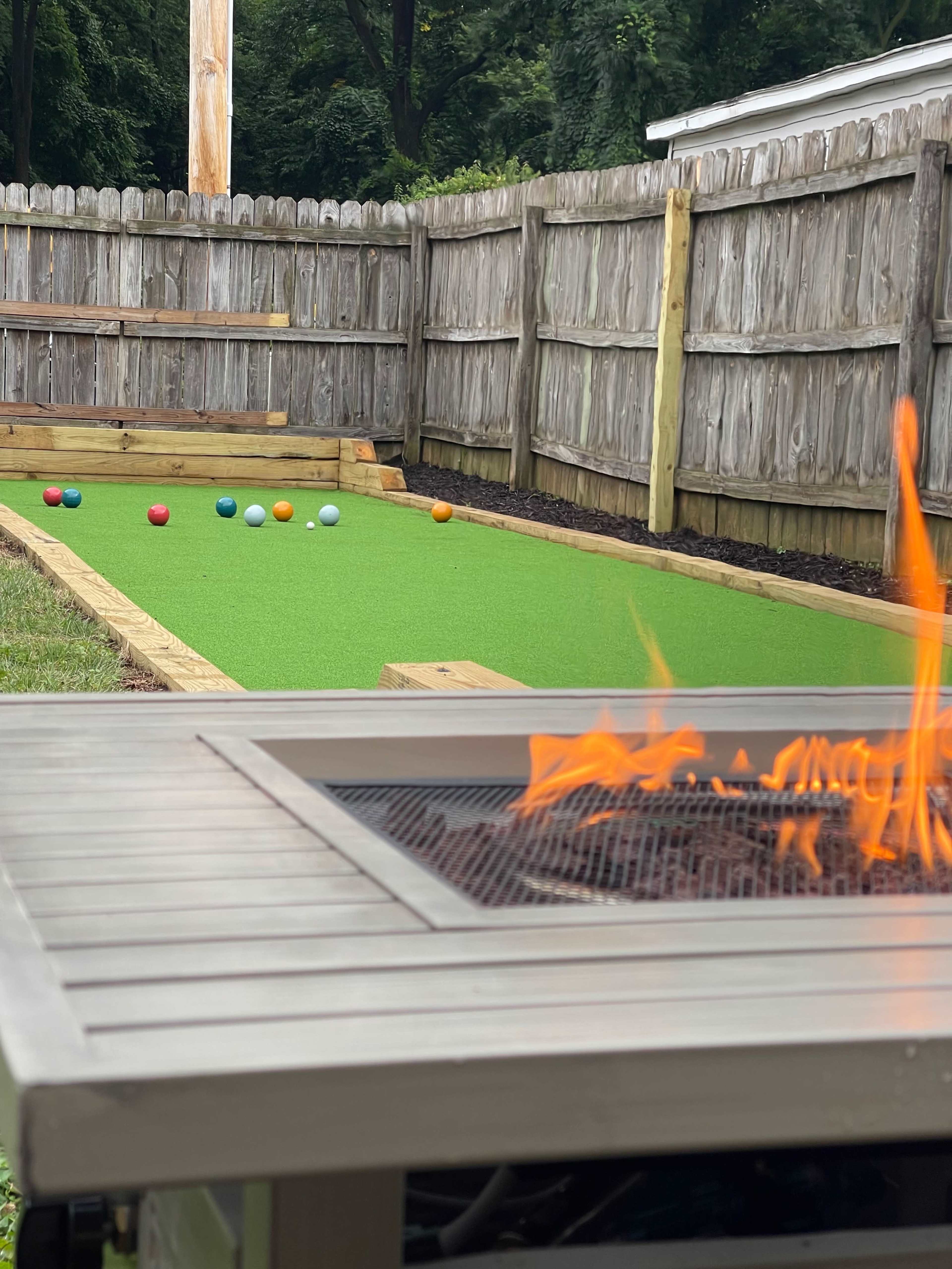 A backyard with a bowling green, featuring colored balls arranged on a synthetic grass surface and a fire pit in the foreground.