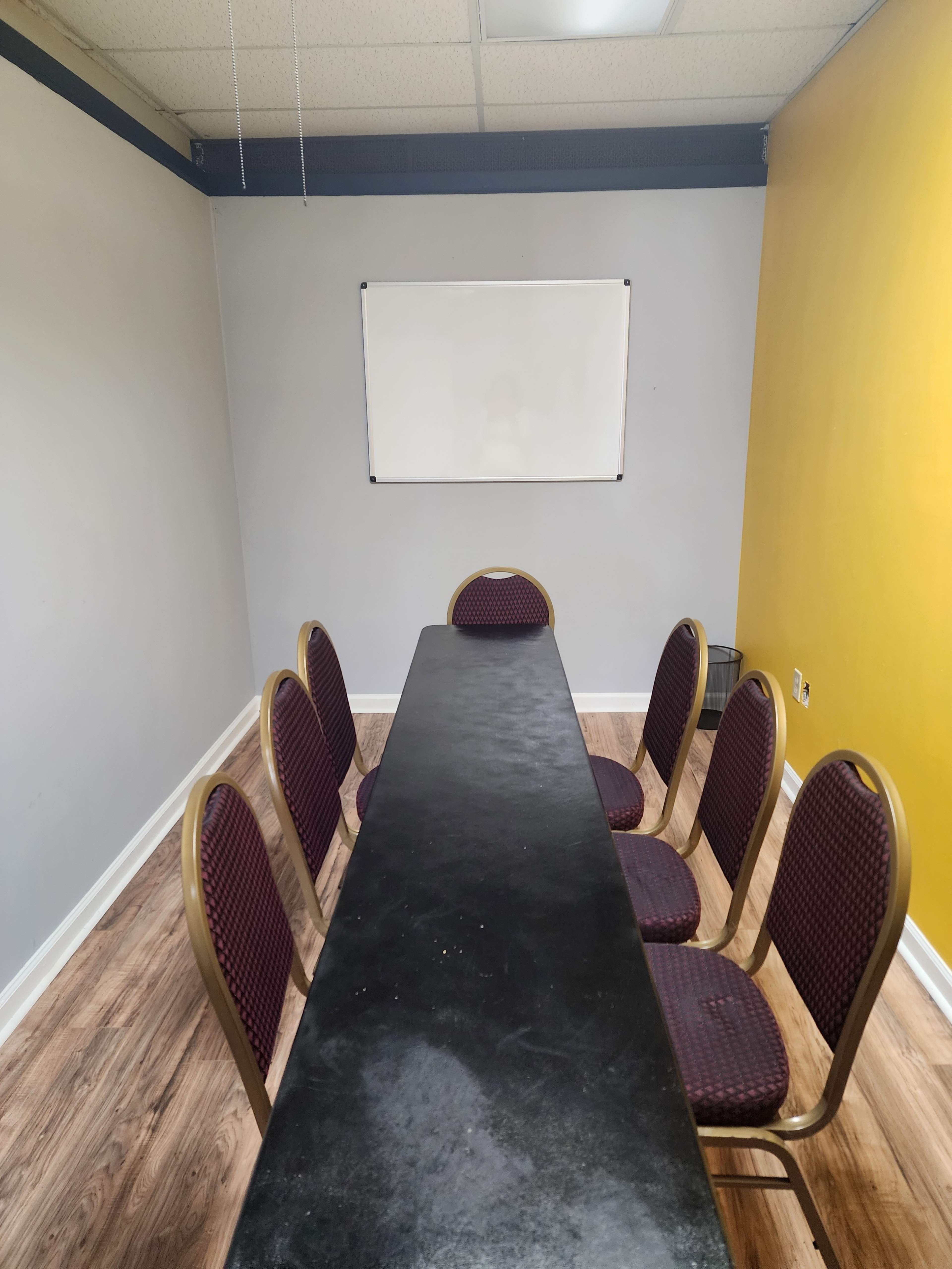A meeting room features a long table with six chairs arranged around it, and a whiteboard mounted on the wall.