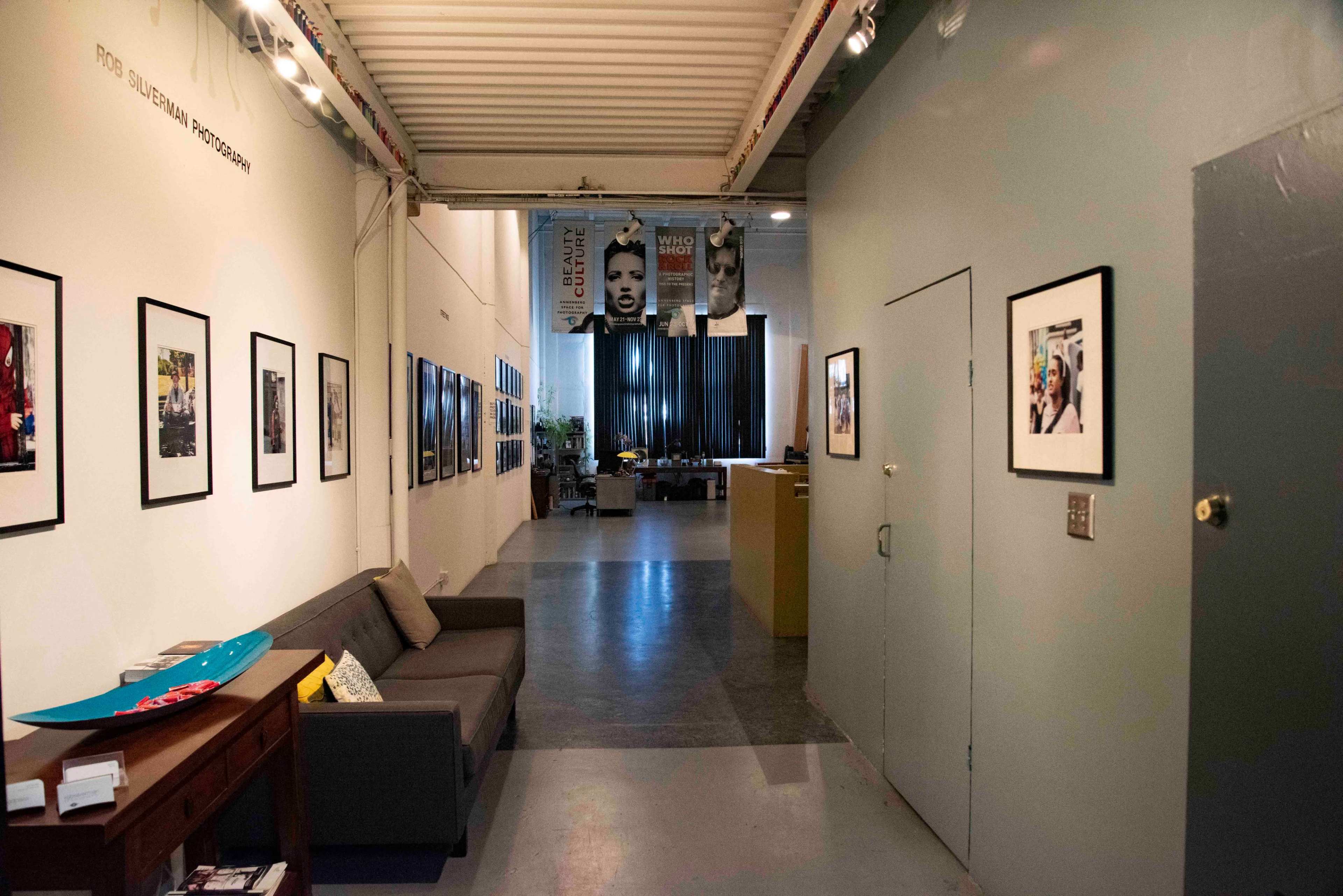 A hall in an art gallery with framed photographs on the wall and a view into a workspace area at the end.