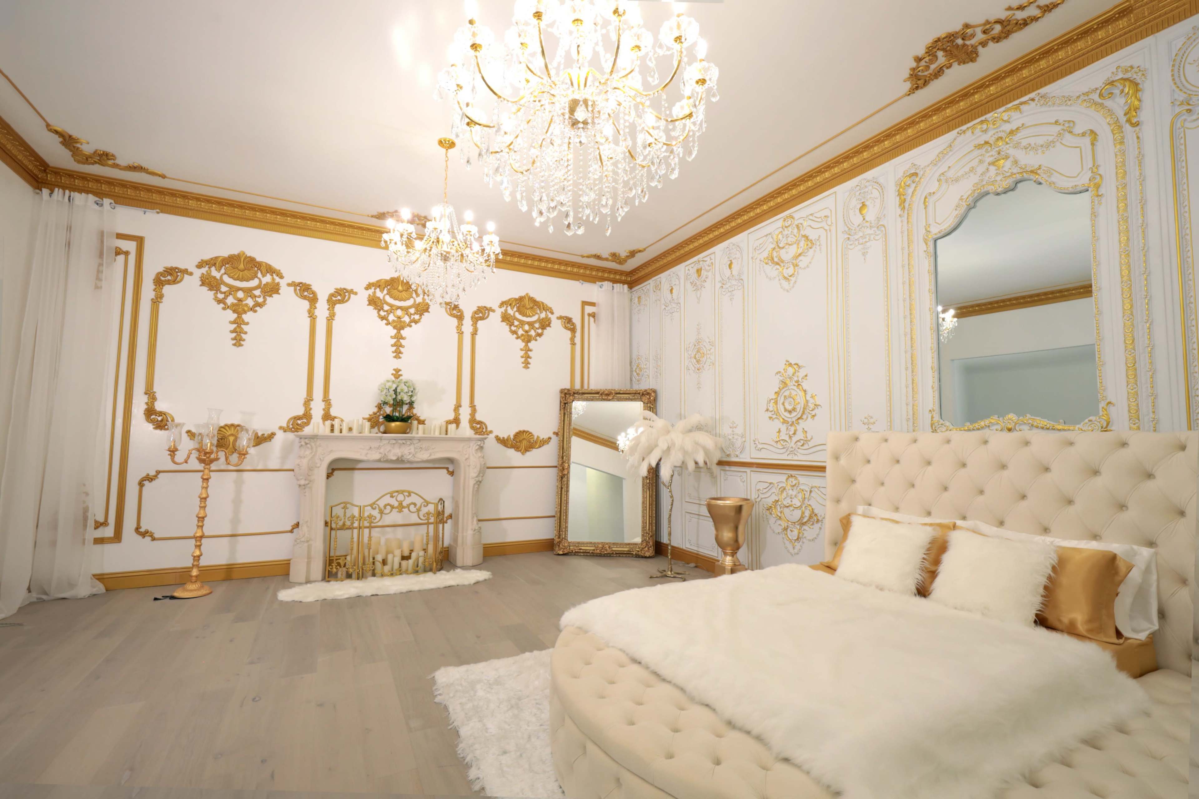 A lavish bedroom with ornate gold accents, a plush bed, a large mirror, and an elegant chandelier.