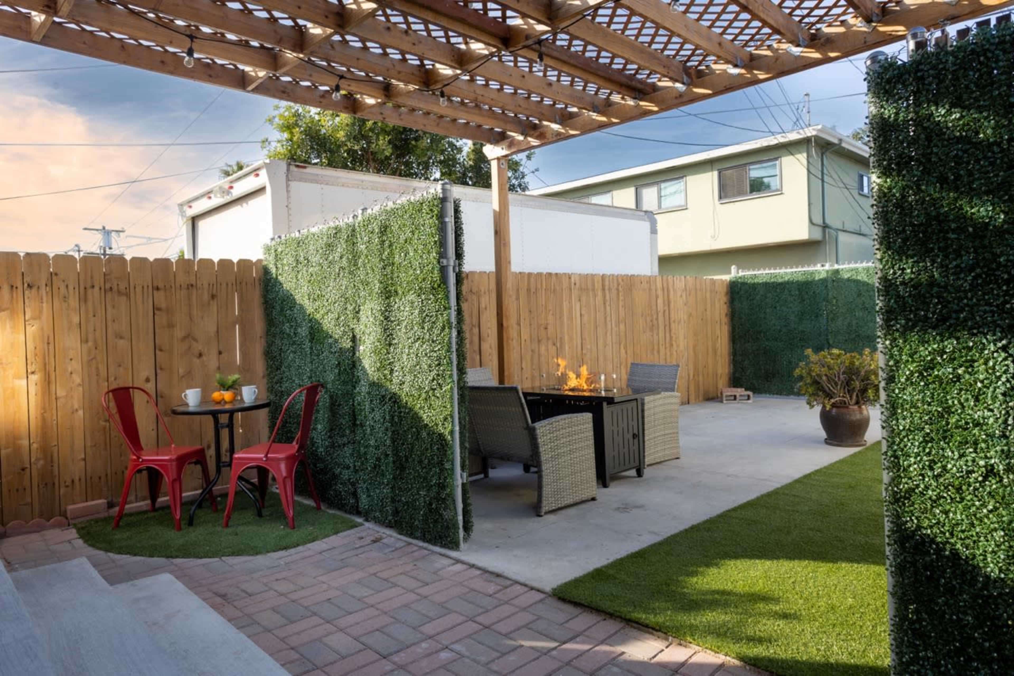 The image shows a patio area enclosed by greenery and a wooden fence, featuring a seating arrangement around a fire pit and a small table with chairs.
