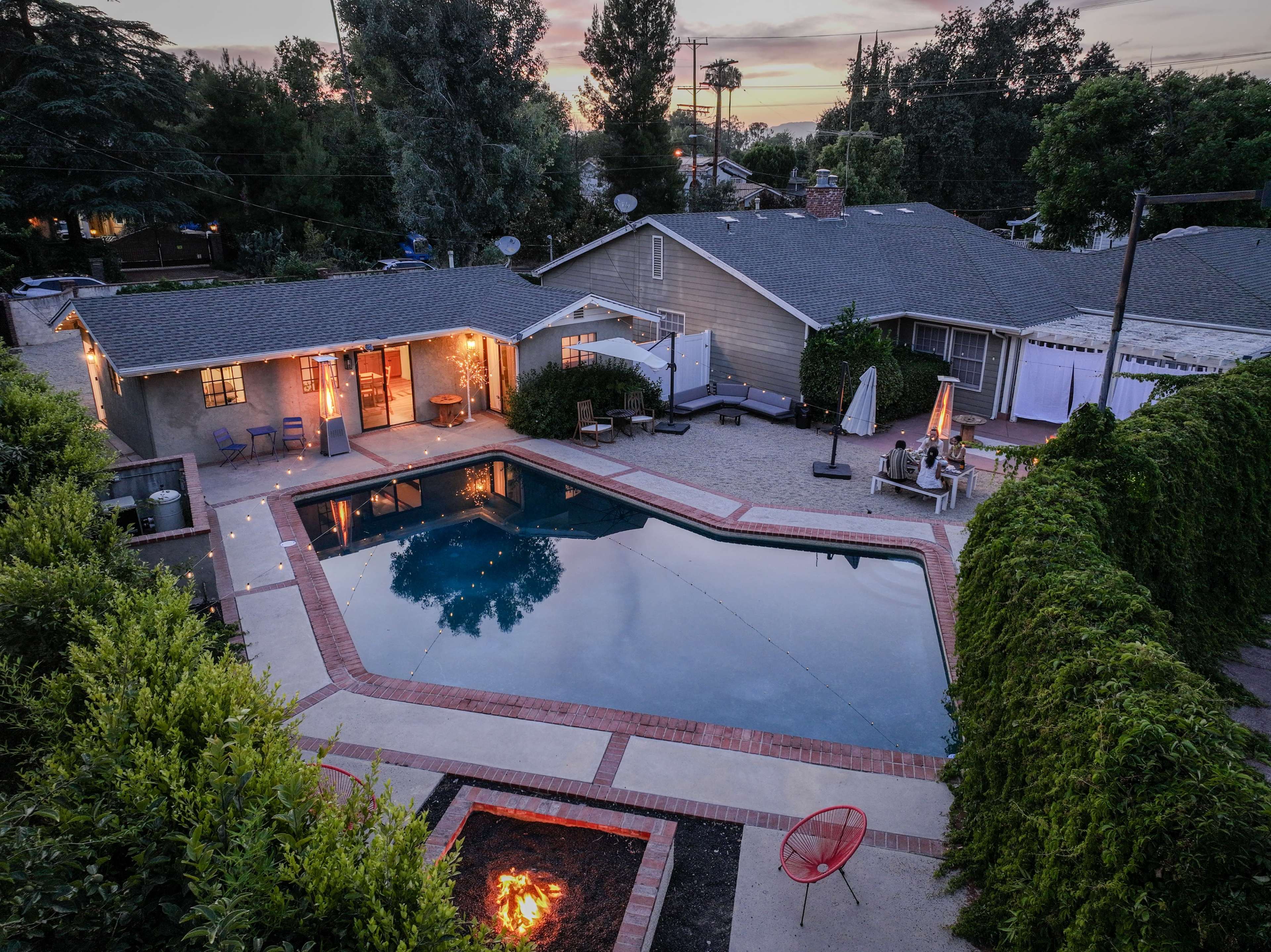 A backyard view features a swimming pool surrounded by a patio and a fire pit, with a house and lush greenery visible in the background.