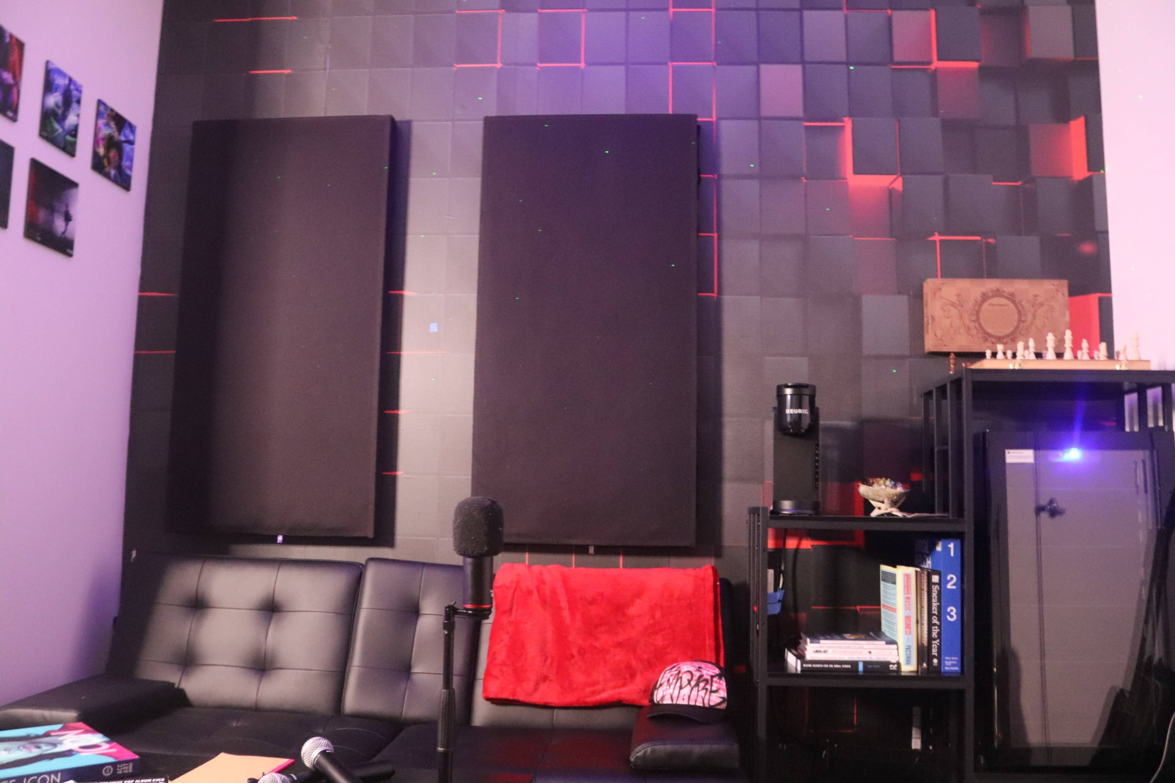 The image shows a modern room with a black seating area, soundproofing panels on the wall, and a bookshelf filled with various items.