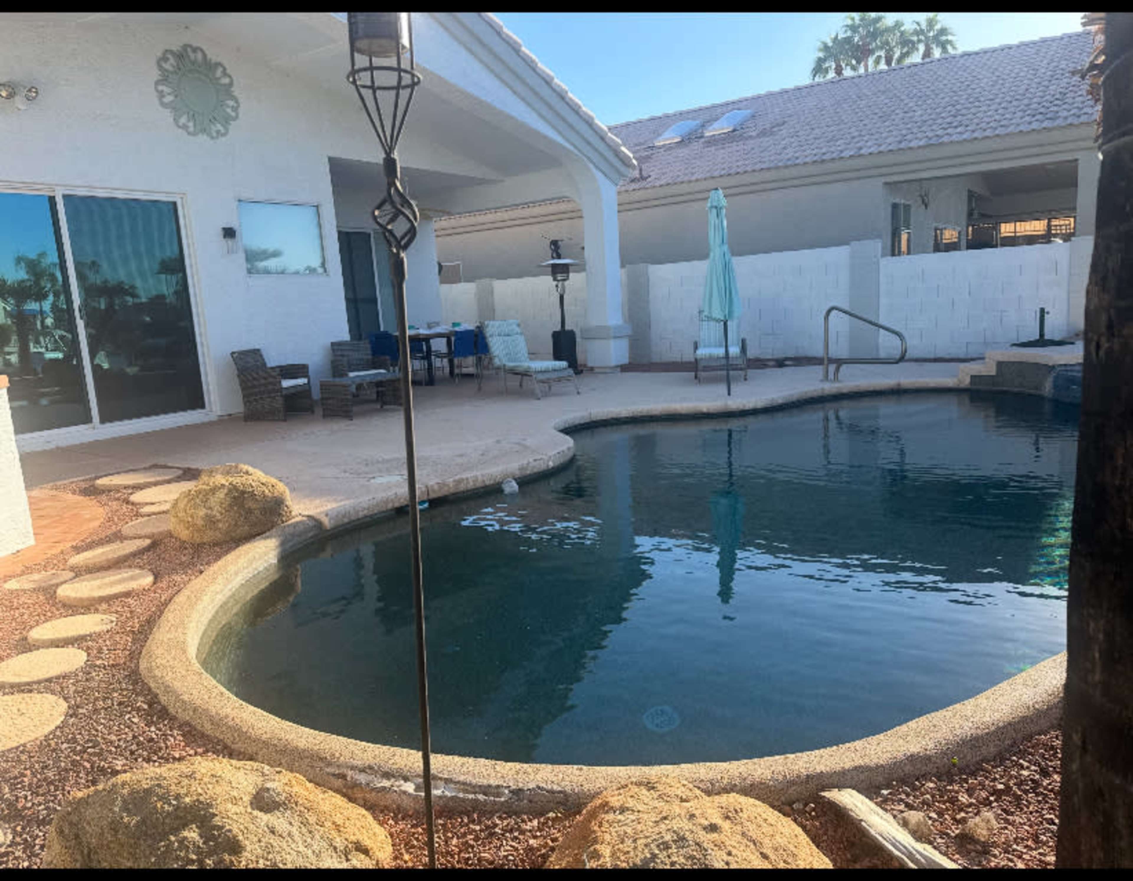 The image shows a backyard with a swimming pool surrounded by stone and gravel, and a shaded seating area in the background.