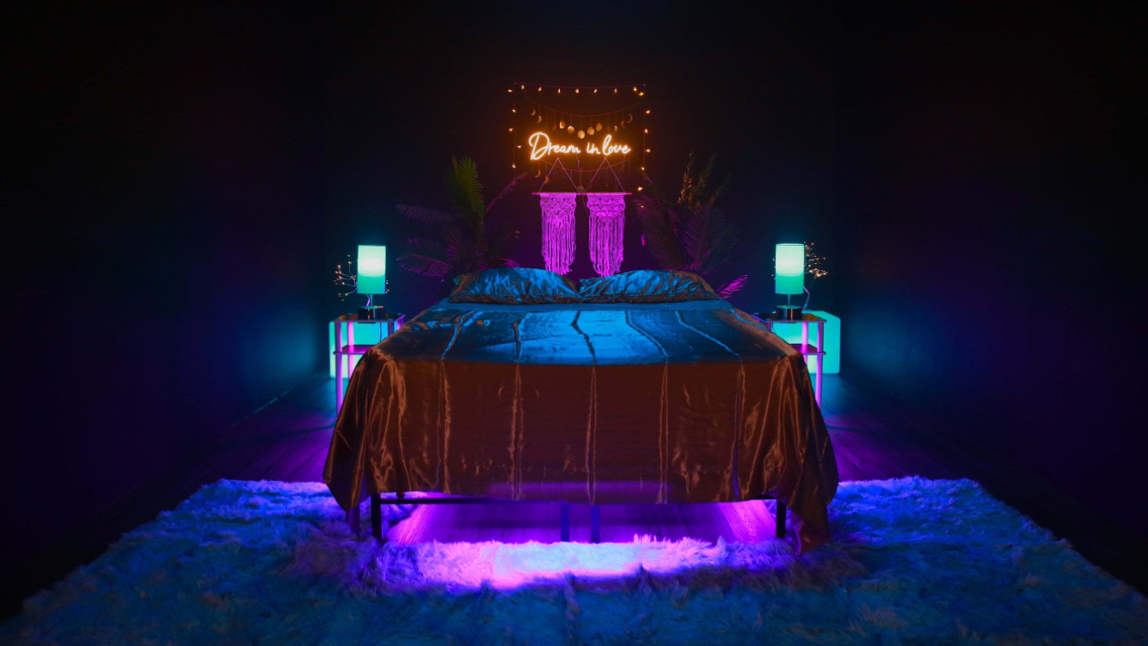 A well-lit bedroom features a bed with a velvet cover, surrounded by colorful lights, plant decorations, and a neon sign above it.