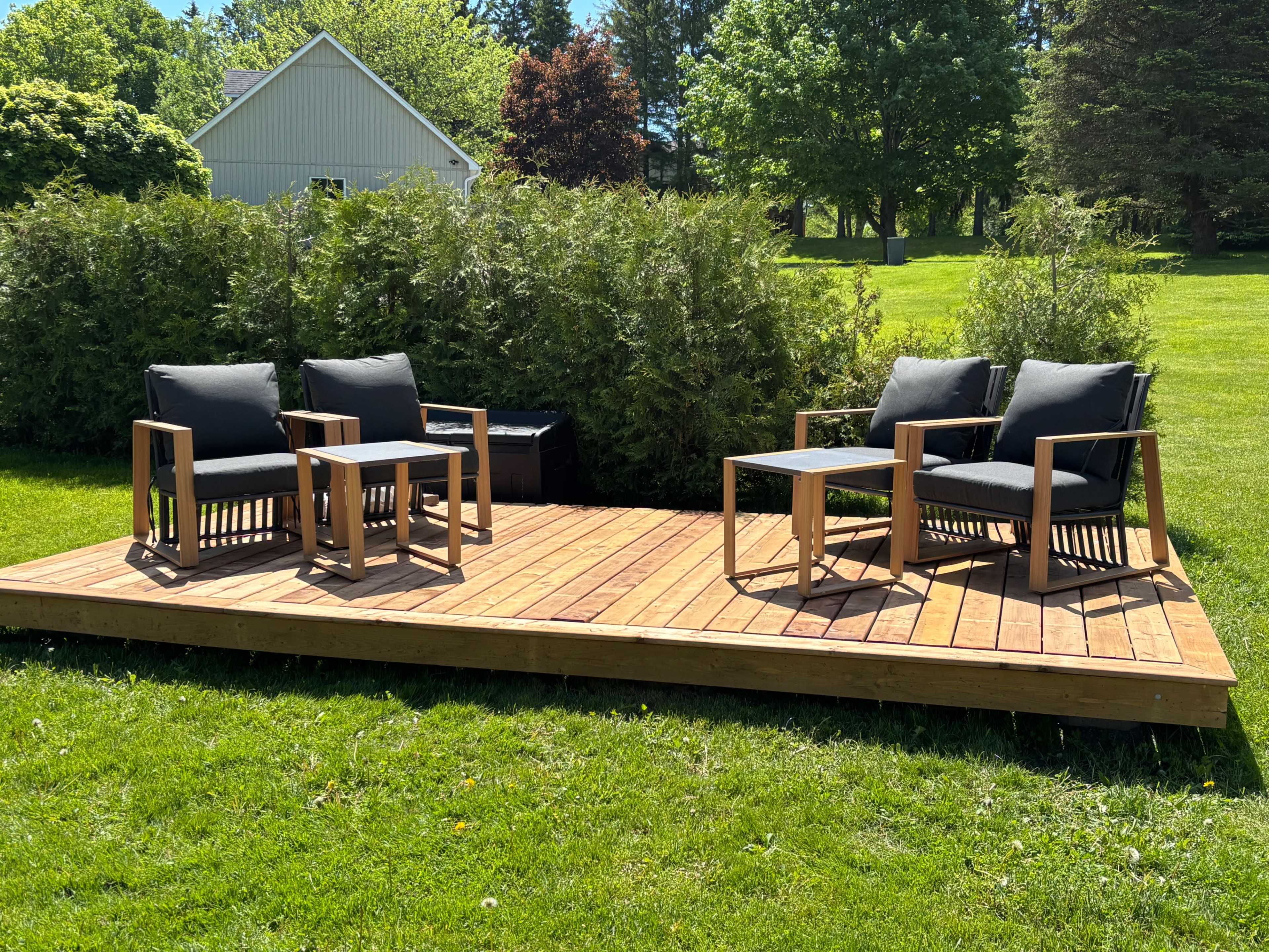 A wooden deck features four black cushioned chairs and two small tables set on a grassy area surrounded by trees.