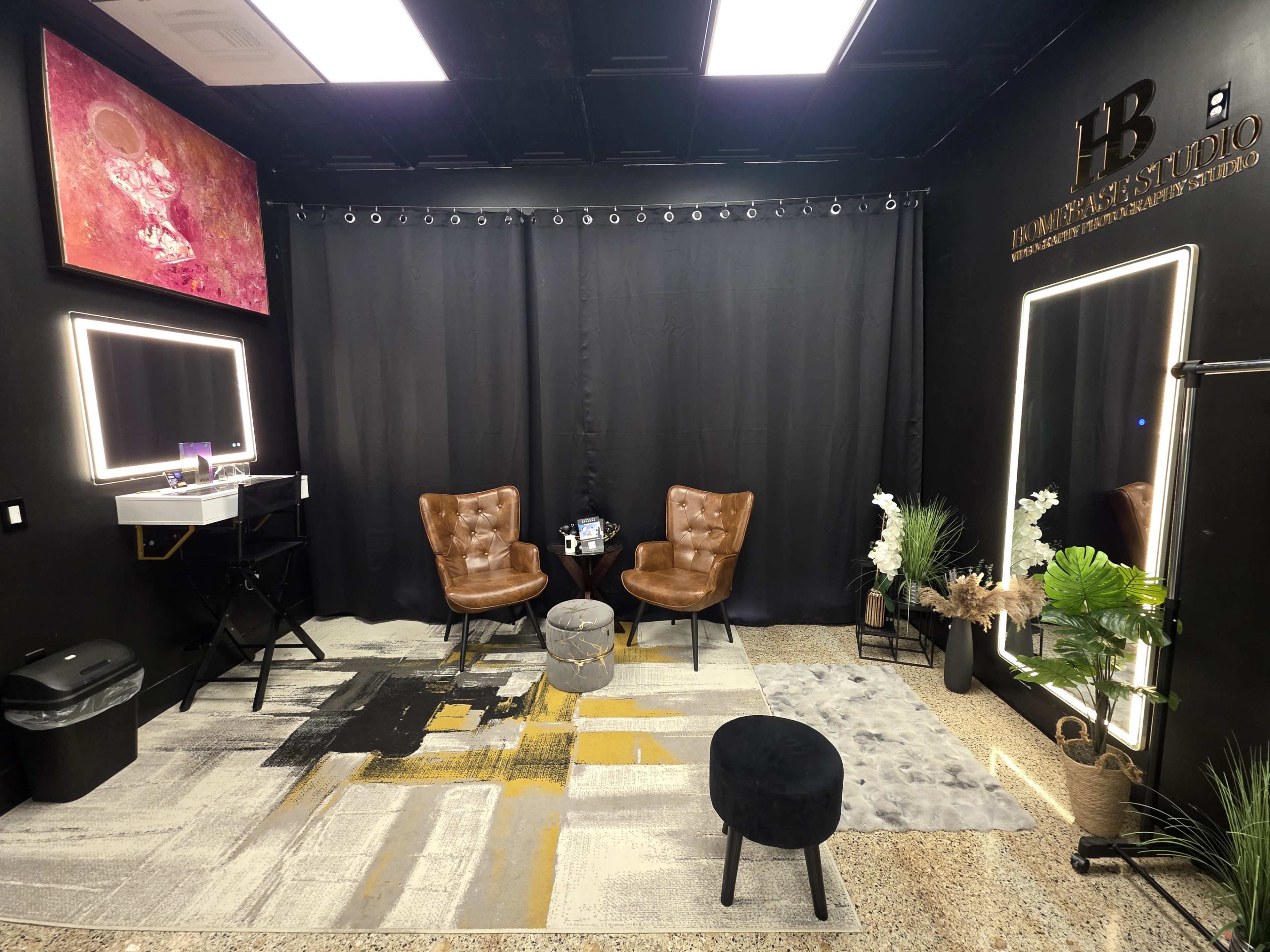 The image shows a modern beauty studio featuring two brown upholstered chairs, a small round table, and a large mirror, all set against a backdrop of black curtains and decorative elements.