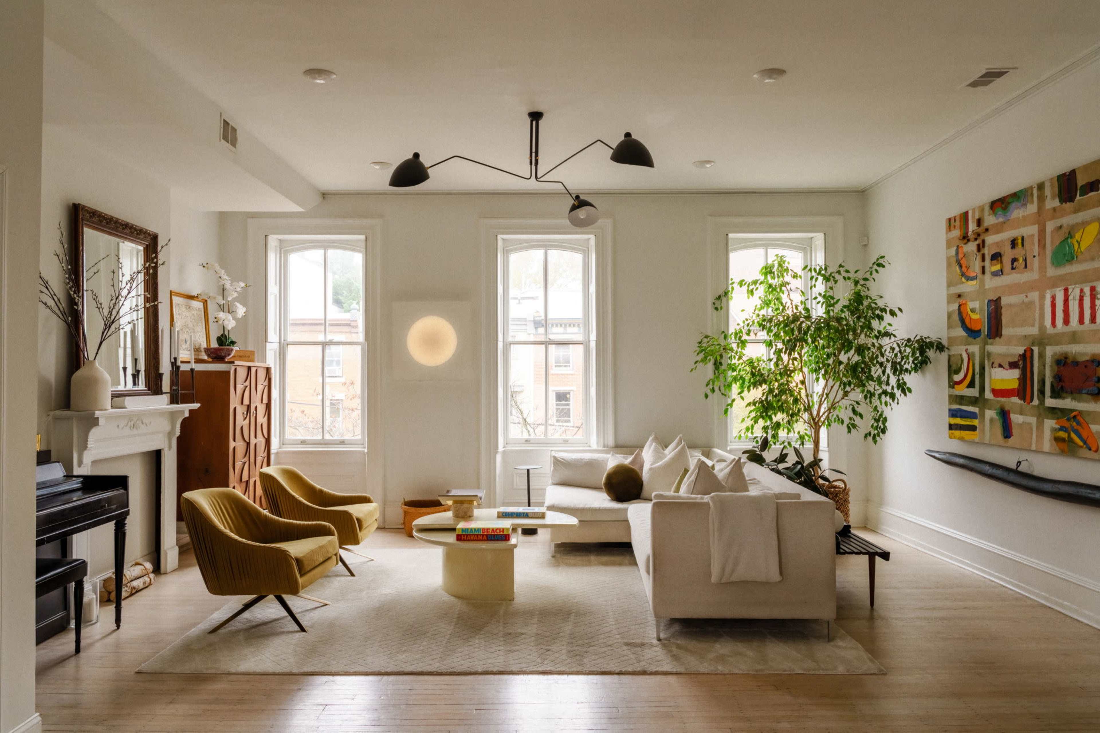 The image shows a bright living room featuring two large windows, a beige sectional sofa, yellow accent chairs, a round coffee table, and a large abstract painting on the wall.