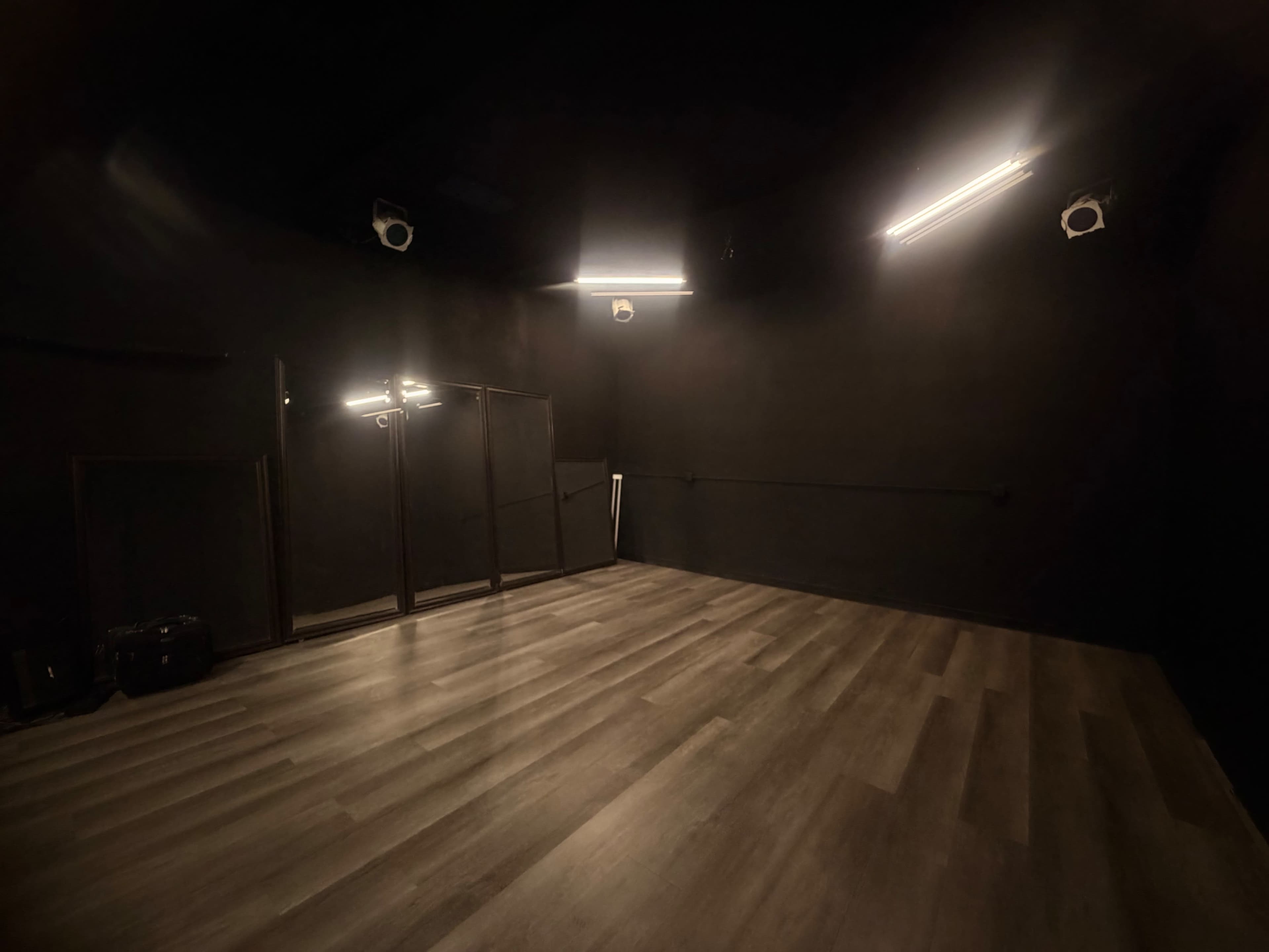 The image shows a dark room with black walls and wooden flooring, featuring a large mirror and overhead lights.
