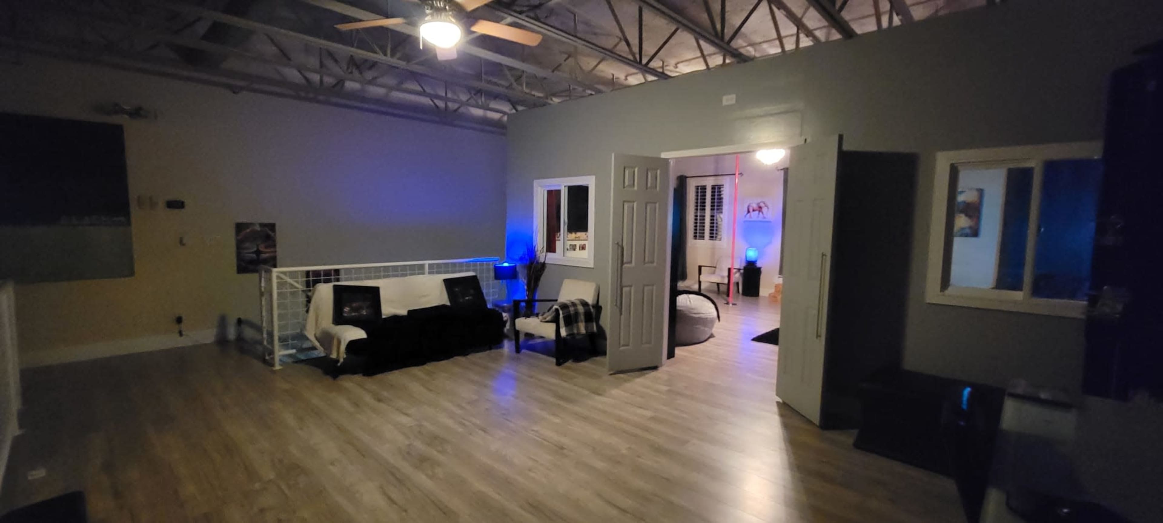 The image shows a spacious room with wooden flooring, containing a sofa, chairs, and a doorway leading to another area illuminated by blue lights.